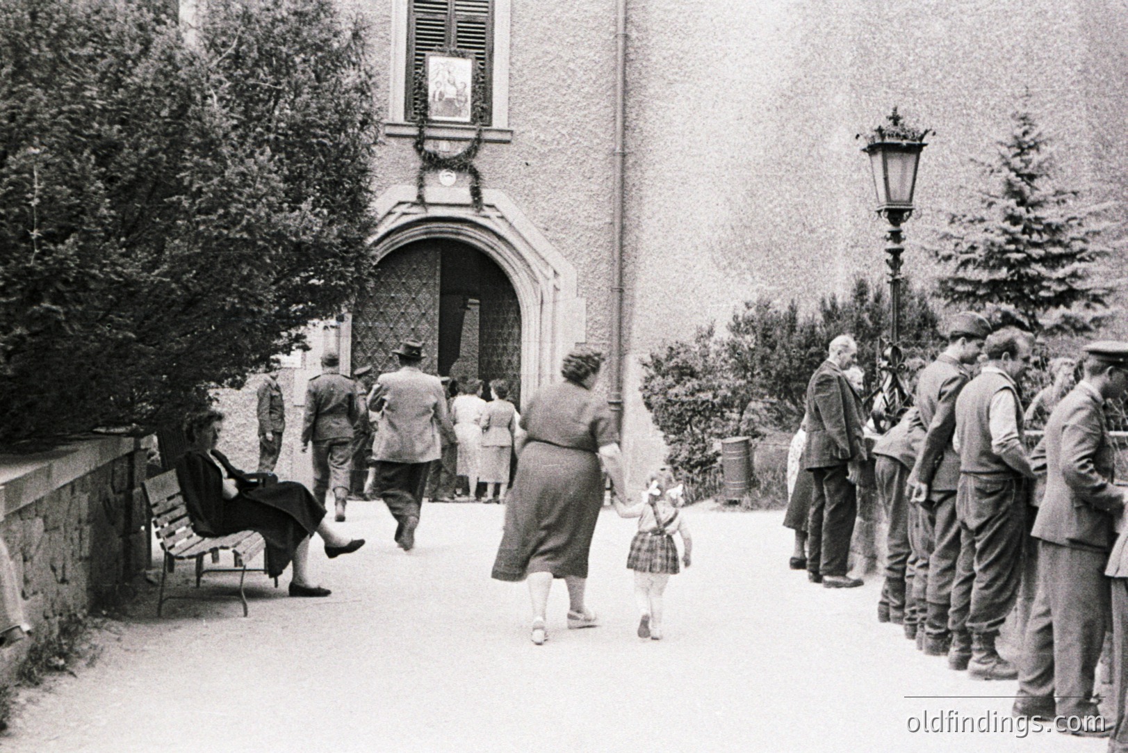 A group of men in military uniforms stand at attention along a paved courtyard, potentially awaiting inspection. A woman and child walk away from the entrance of a grand building, framed by greenery. A seated woman observes. Appears to be post-war Europe, potentially 1950s.