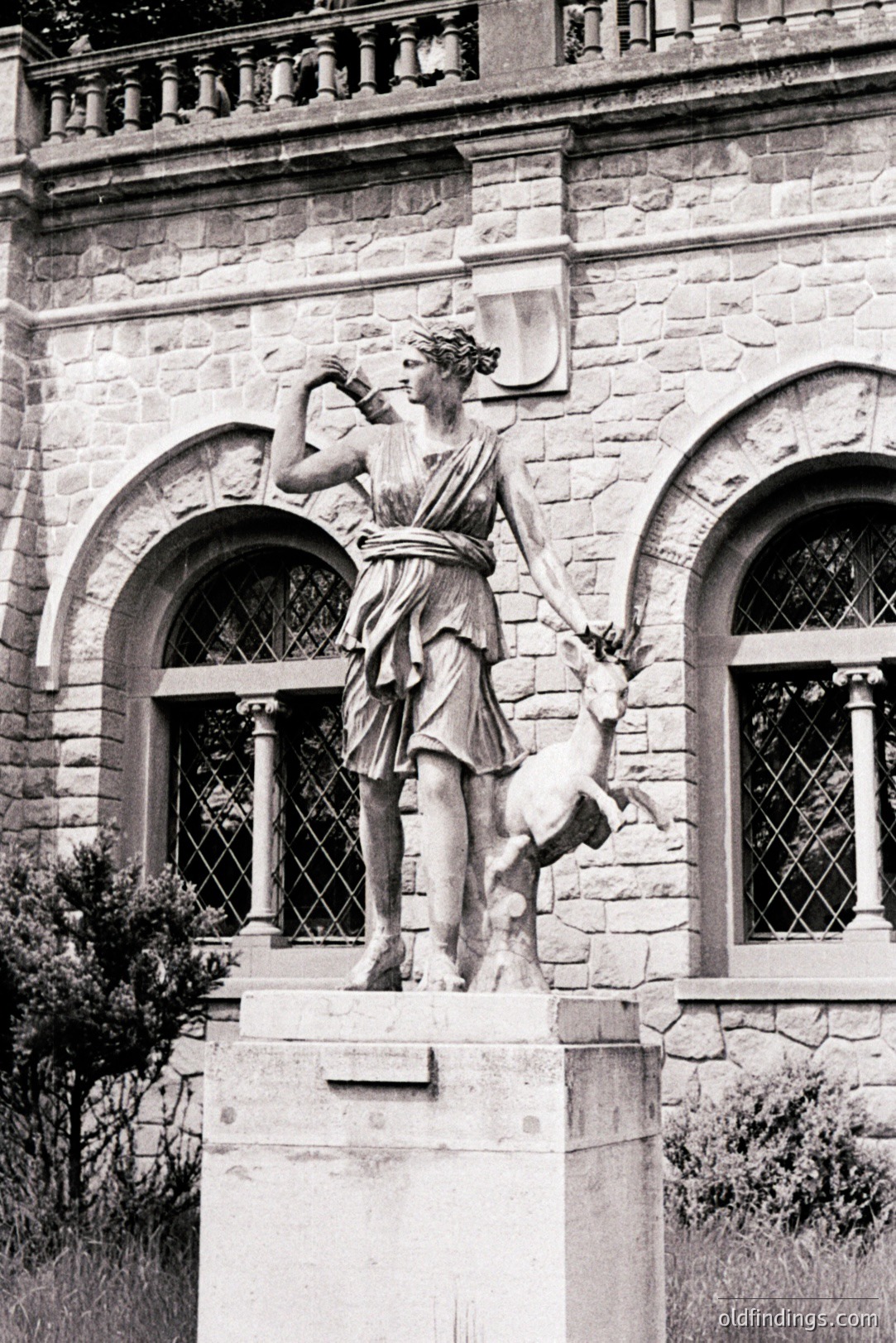 Elegant, classical sculpture of a woman (Diana/Artemis?) with a fawn. Stone carving is atop a pedestal, positioned before a stately building with arched windows and detailed stonework. Likely late 19th or early 20th century. A reference for landscape design & neoclassical art.