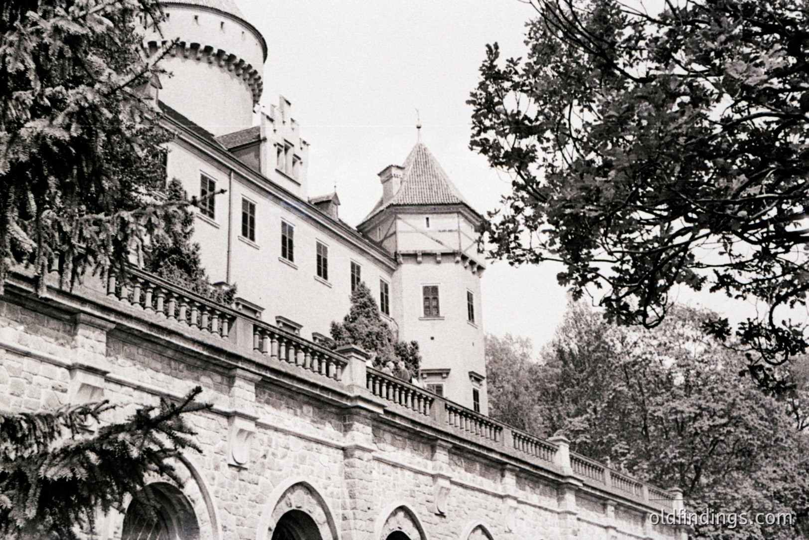 A stately stone manor with crenelated towers rises above an arched walkway. The building displays arched windows and a detailed balcony railing. Likely an alpine estate, demonstrating late 19th or early 20th century architectural influences. Potential stock photography asset for design or historical research.