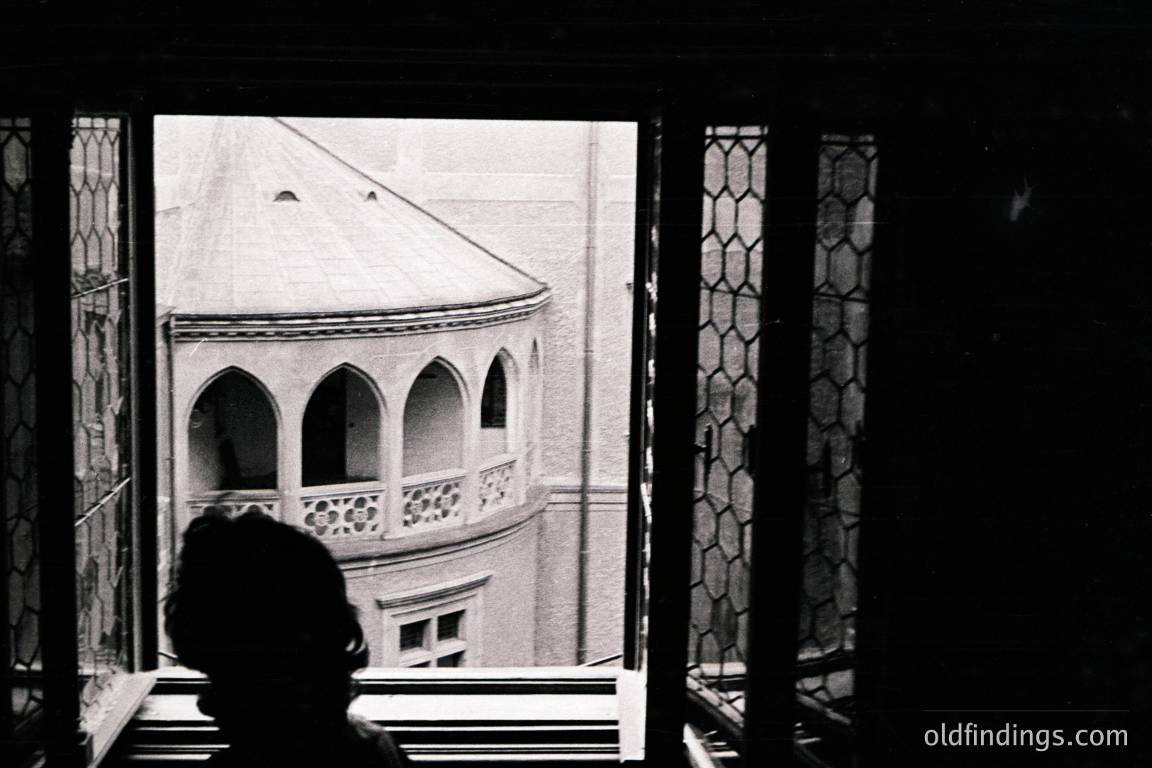 Framed view through a window reveals a building with arched balconies, showcasing architectural details. A figure's silhouette is visible, gazing out. Likely a travel or architectural study, suggestive of a European location. Appears to be a vintage photograph, possibly 1950s-1970s.