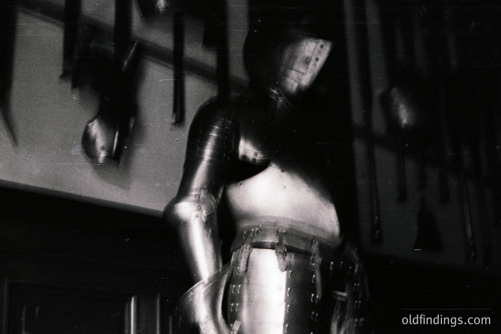 Detailed, partial view of a knight in full plate armor, photographed in a museum setting. The armor is highly reflective, showing highlights and shadows. Weapons are visible on the wall behind. Likely mid-20th century museum documentation. Commercial value as stock or reference image for historical designs.
