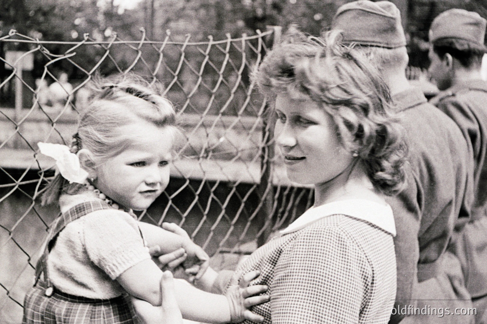 A young girl in a plaid dress and ribboned hair reaches through a chain-link fence towards a smiling woman. Two uniformed soldiers stand behind, suggesting a military presence or restricted access. Likely post-war Europe, 1940s-1950s. Poignant image of connection amidst separation.