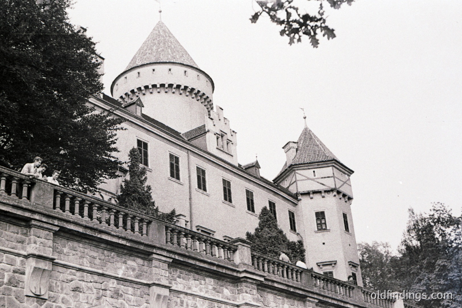 A view of a stone castle with multiple turrets and a balcony, partially obscured by foliage. Two figures are visible on the balcony, observing the view. Likely a European estate, possibly mid-20th century. Architecture suggests historical influence. The image has potential for design reference or historical study.