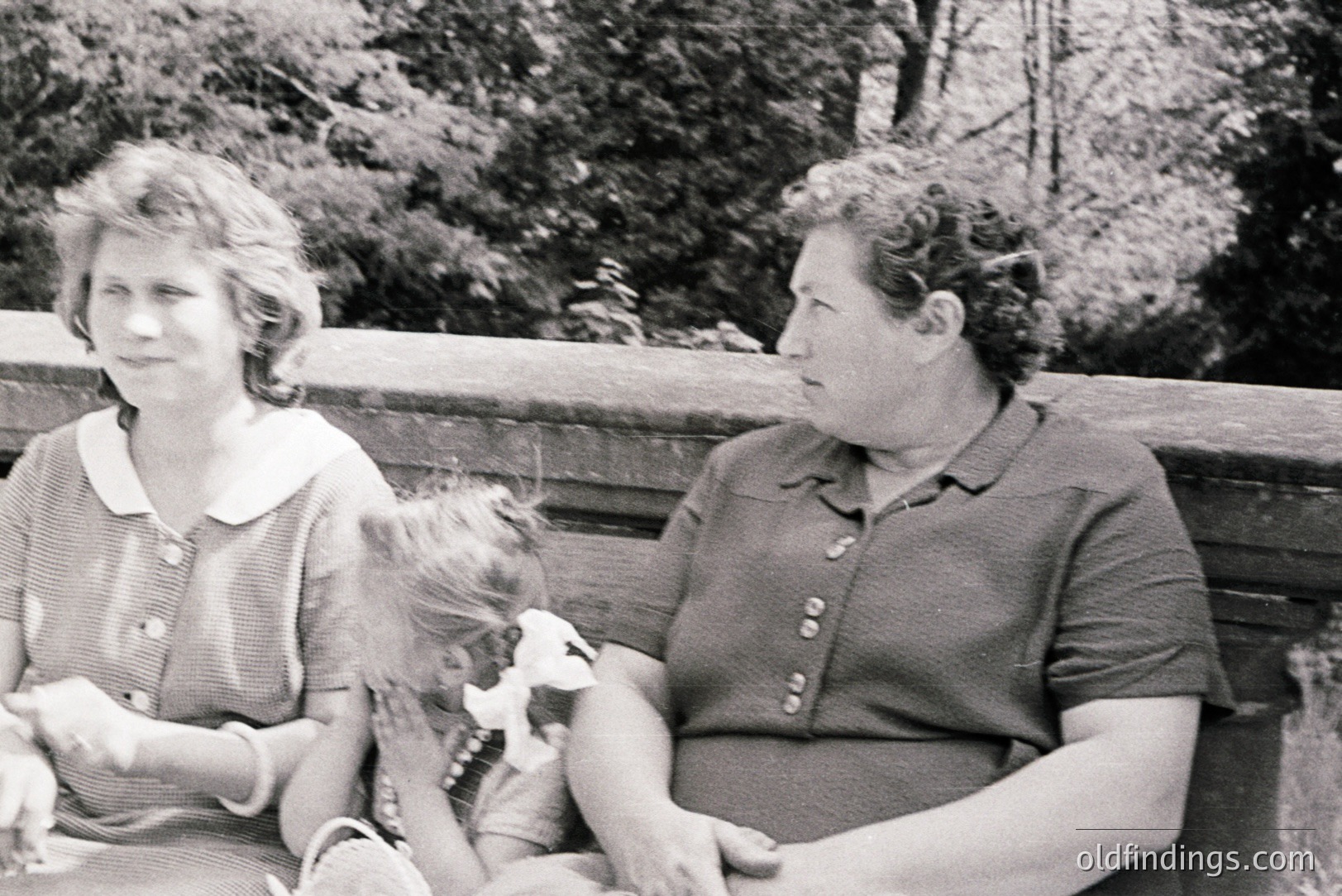 A mother and young girl sit on a wooden bench, likely in a park or garden. The girl holds a stuffed toy. The mother wears a short-sleeved dress; both appear comfortable and relaxed. Appears to be a snapshot from the 1960s. Good candidate for family portraiture or design reference.
