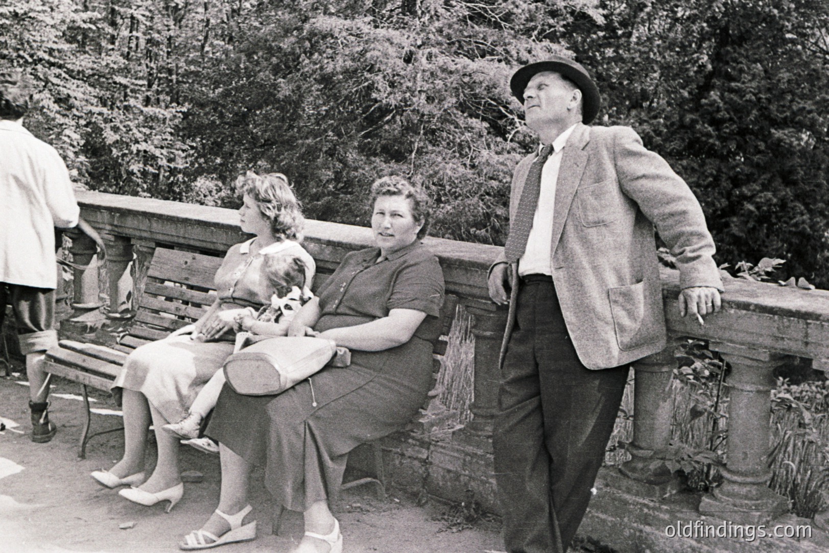 A man in a tweed suit and hat observes two women seated on a bench. The women are dressed in 1950s style attire and one holds a rolled blanket. Stone architecture and foliage suggest a park setting. Classic mid-century portraiture.