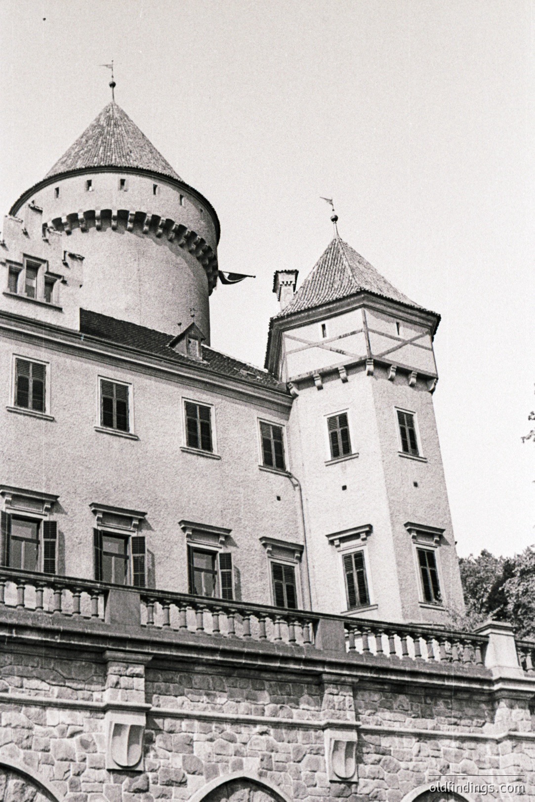 Exterior view of a castle with a central round tower and a square tower with a steeply pitched roof, both topped with decorative finials. Stone balustrade runs along the upper level. Likely 19th-century European architecture, possibly a chateau or manor. Notable for its symmetrical design.