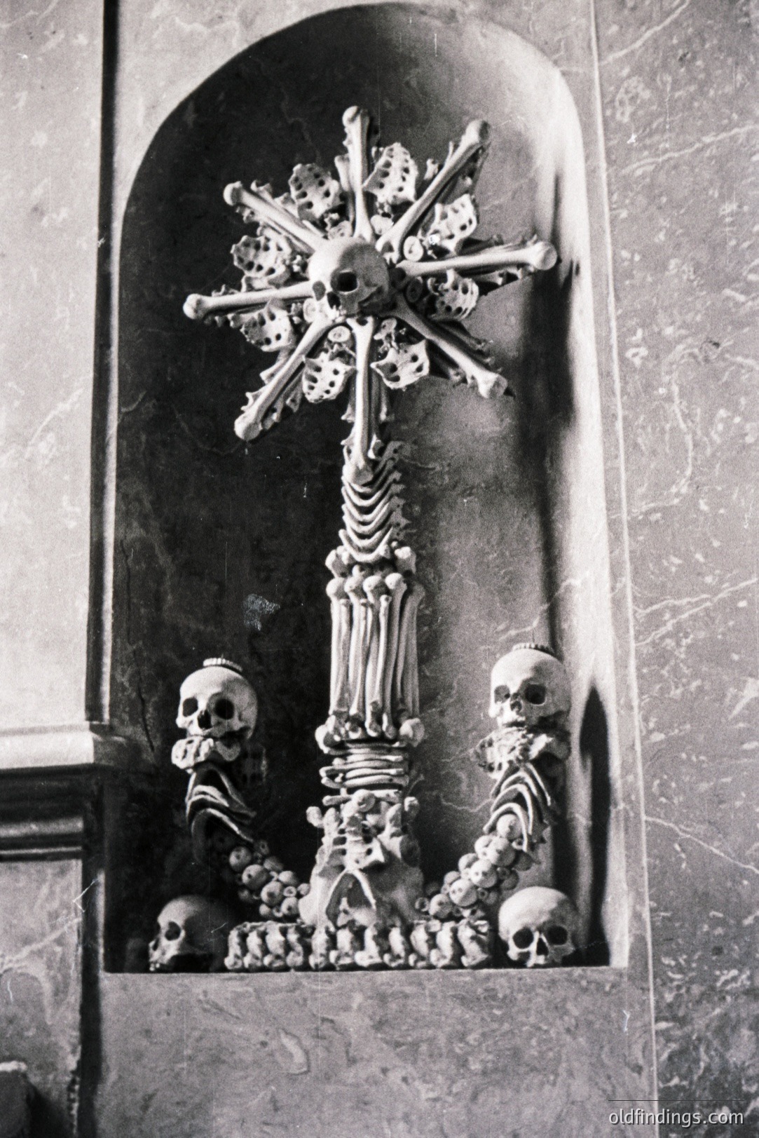 A striking, monochromatic depiction of a skeletal cross within a niche. Human skulls and bones meticulously arranged form the cross’s structure and base. The artistic piece represents mortality and religious iconography. Likely dating to the 18th or 19th century, potentially found in a European religious setting.