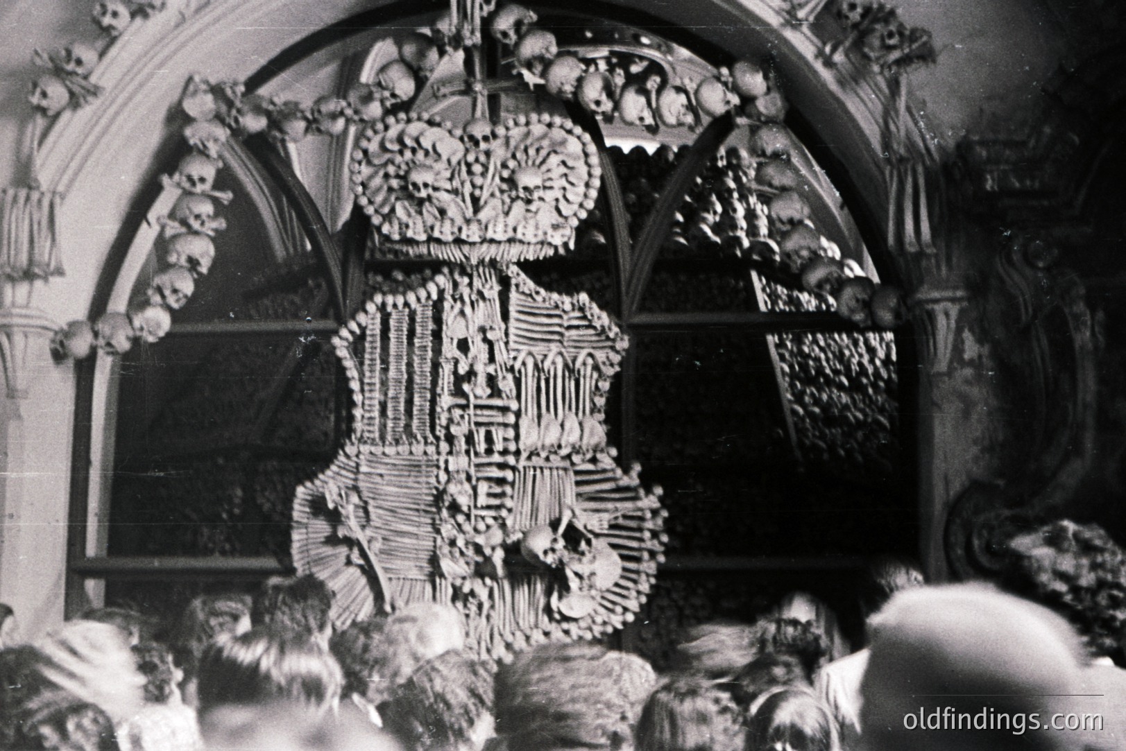 A complex, intricately crafted artwork composed entirely of human bones and skulls dominates the view. Likely a devotional piece within a church, it displays heraldic motifs and ornate detailing. Spectators are visible in the foreground, blurred. Likely 18th or 19th century, Europe.