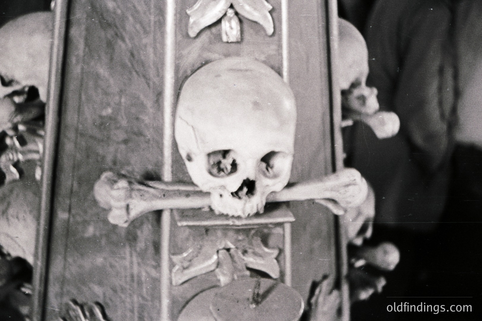 A macabre display featuring a human skull crossed with tibias, forming a "memento mori" symbol. Likely a museum exhibit or theatrical prop. The stark black and white tones suggest a vintage photograph, possibly from the mid-20th century. The background appears to be a carved wooden panel.