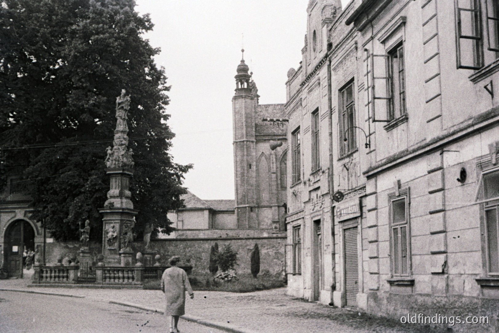 Formal, monochromatic image shows a 1960s European town square. A woman in a coat walks toward an ornate stone column topped with a statue. Background features a walled garden, a brick building with a steeple, and a building with shuttered windows. Captures architectural detail & local life.
