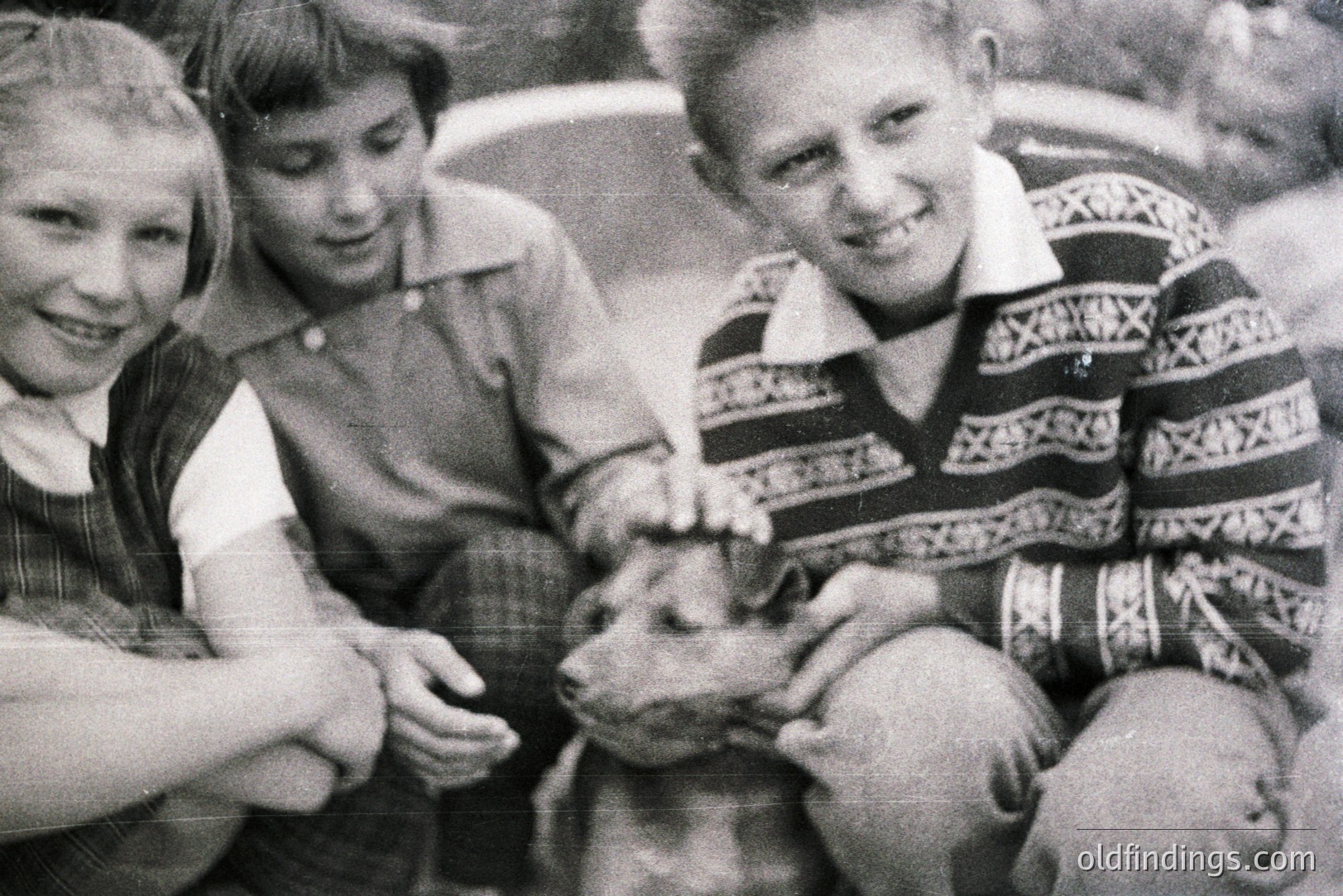 Three children pose with a young goat. Two boys flank a girl in what appears to be a garden setting. The boy on the right wears a patterned sweater, indicative of mid-century style. Grainy black and white image likely from the 1960s or 70s. Possible family snapshot.