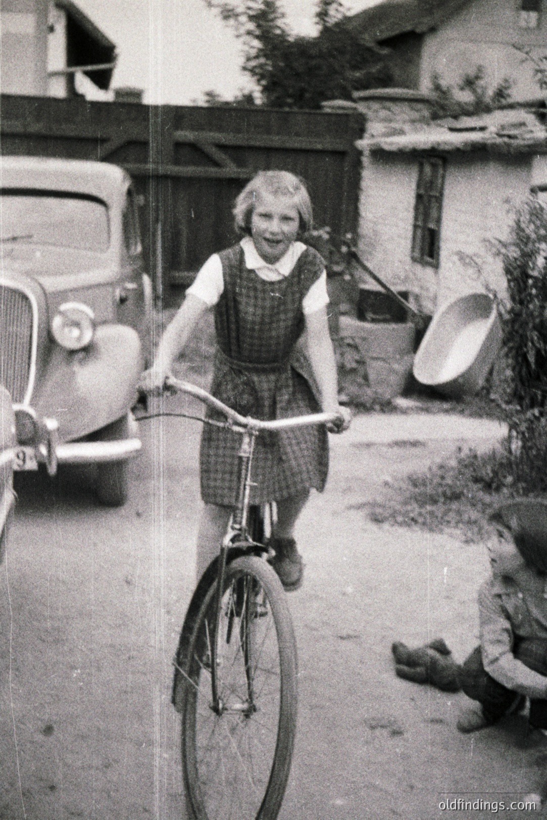 Young girl on a vintage bicycle, captured in motion. Classic 1950s car visible in the background along with simple housing/outbuildings. Likely a backyard scene. The photograph holds nostalgic appeal; would be strong for design or historical research.