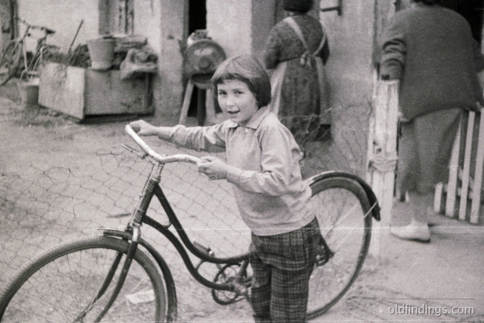 A young boy smiles while holding a vintage bicycle, standing outside a rural home. The home's exterior shows weathered siding and a simple porch. A woman in a long dress stands partially visible in the doorway. Likely a candid snapshot from the mid-20th century.