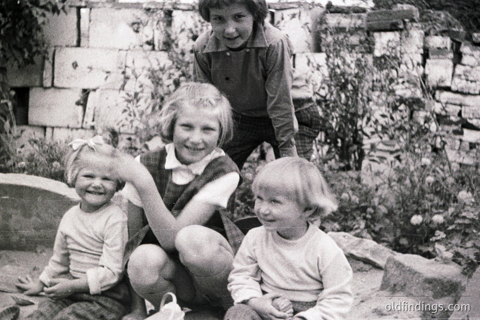 Four children pose outdoors; three seated, one standing, amidst a stone wall and foliage. The style of dress, including overalls and short haircuts, suggests a mid-20th century, likely 1960s, setting. Candid portrait captures childhood playfulness. Image suitable for family history research or vintage design.