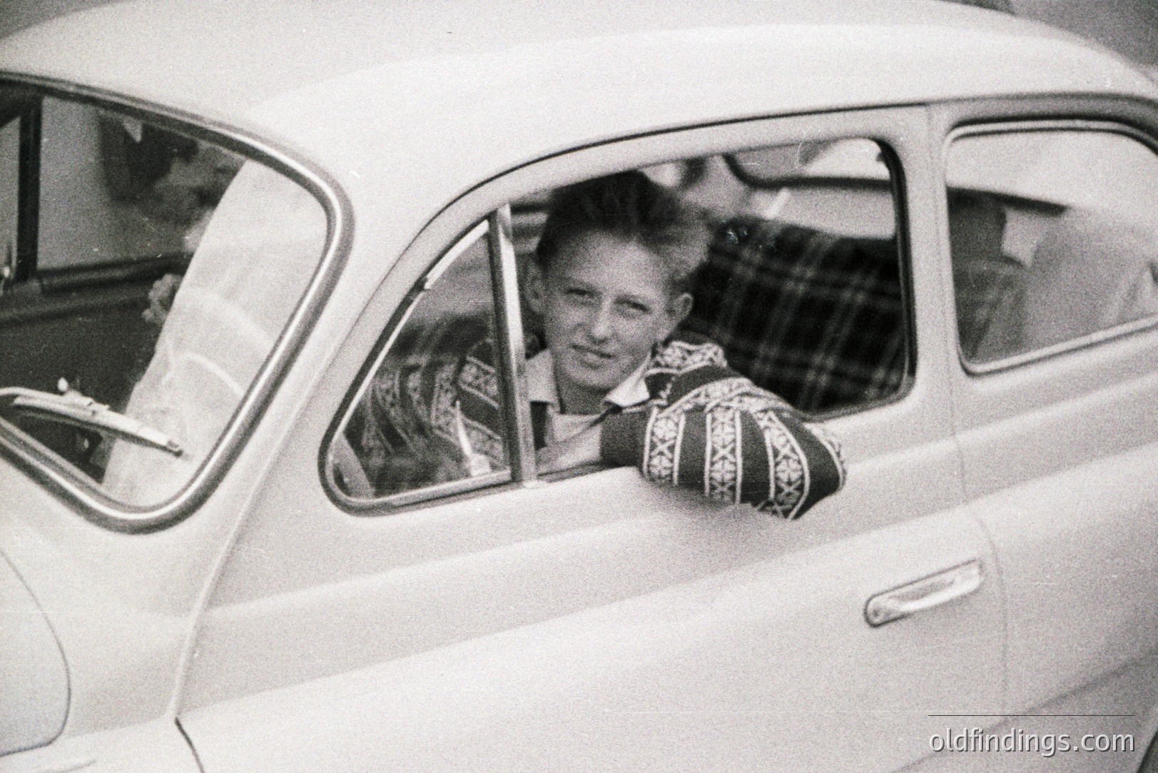 Young man leans out of a vintage Volkswagen Beetle, likely 1960s, exhibiting a casual, relaxed pose. Patterned sweater & short hair suggest a youthful, informal style. Car's design indicates mid-century automotive aesthetics. A moment captured in everyday life.