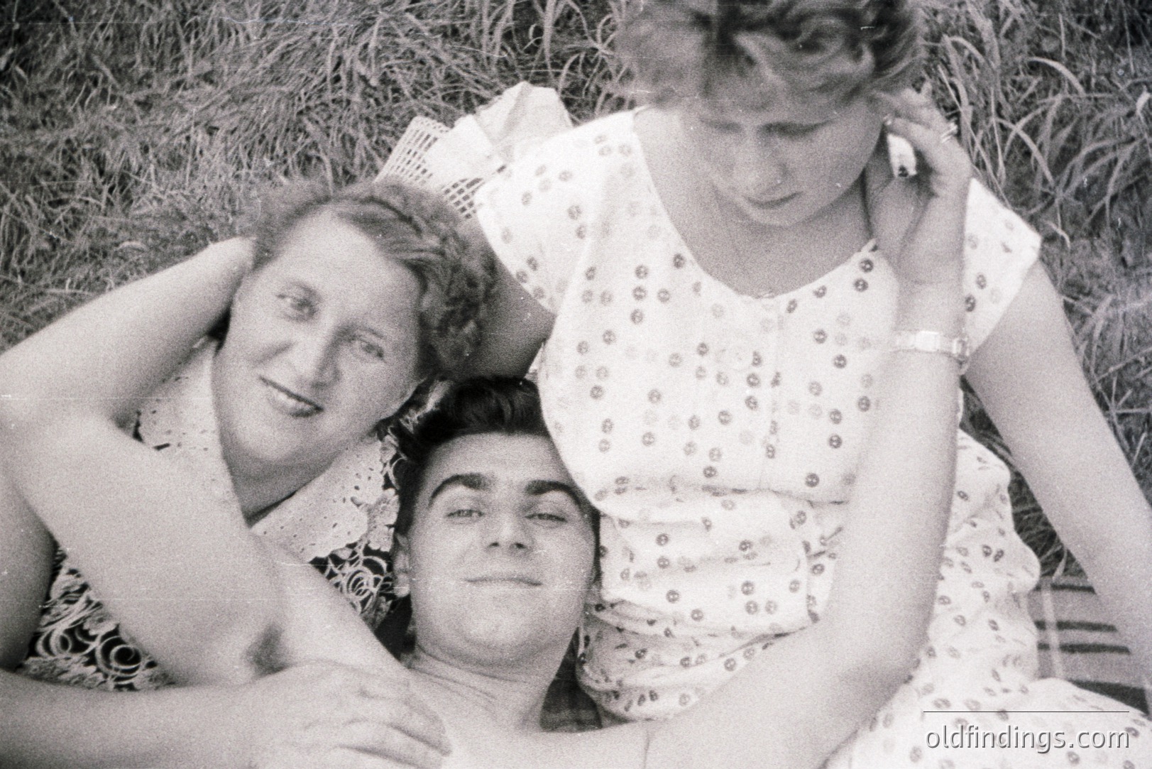 A black and white candid photo depicts a man lying on grass embraced by two women. All three appear relaxed and smiling. The women wear patterned dresses & one has a wristwatch. Likely a 1960s or 70s snapshot, indicative of casual portraiture. A charming, domestic scene.