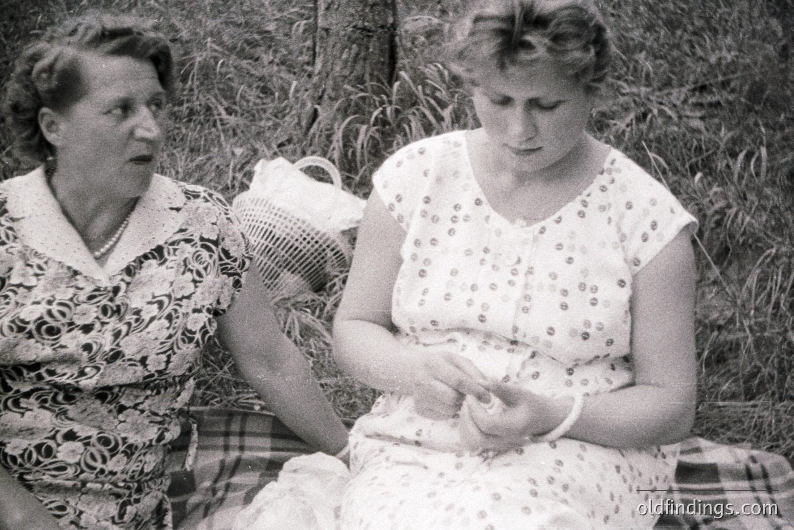 Two women seated outdoors on a picnic blanket, likely in a garden setting. The older woman wears a floral print dress and a necklace. The younger woman, seated closer, examines her hand. A woven basket sits nearby. Appears to be a candid moment, circa 1950s-1960s.