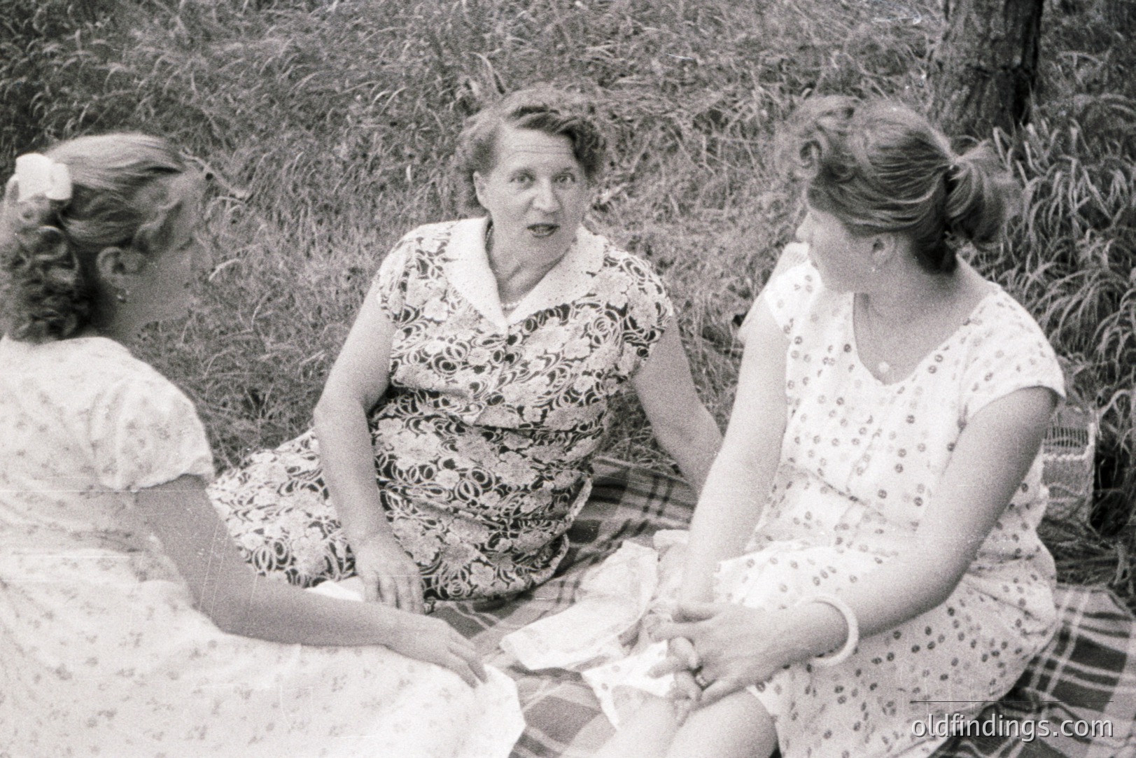 A black and white image shows three women seated on a picnic blanket amidst tall grass. The central woman, likely a mother, appears to be speaking animatedly. All wear floral print dresses typical of the 1950s. The composition suggests a candid, informal moment.
