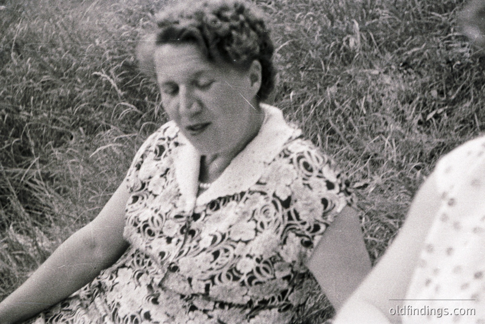 Close-up, black and white image of a woman seated in tall grass. She wears a short-sleeved, patterned dress with a Peter Pan collar. Hair styled in curls. Partial view of another person is visible at right. Likely a casual outdoor portrait.