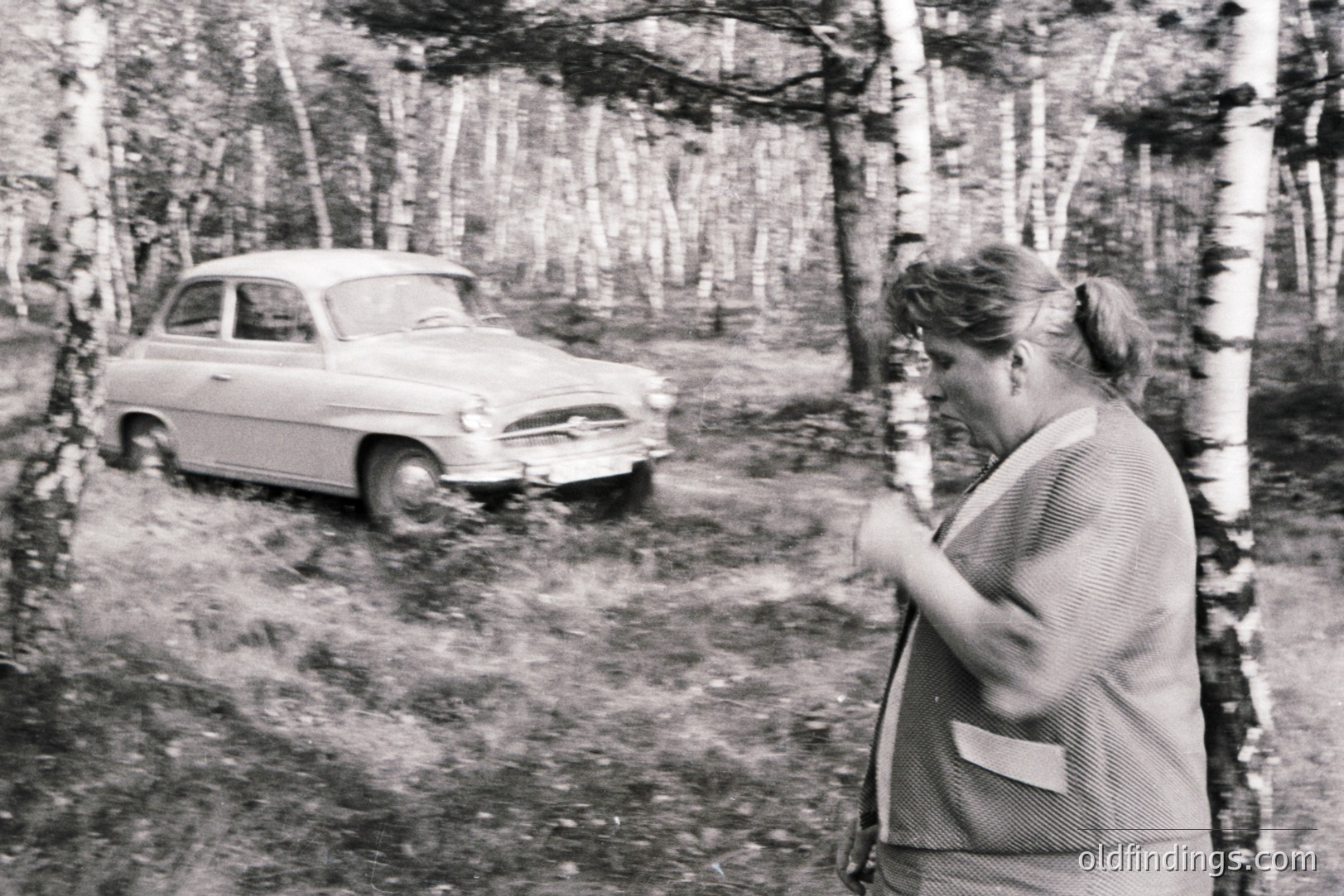 A woman in a patterned suit stands facing away from the camera in a birch forest, alongside a parked, vintage sedan. Appears to be an candid moment, possibly travel or leisure. The style suggests a mid-20th century aesthetic, likely 1950s or 60s. Documentary or personal archive image.