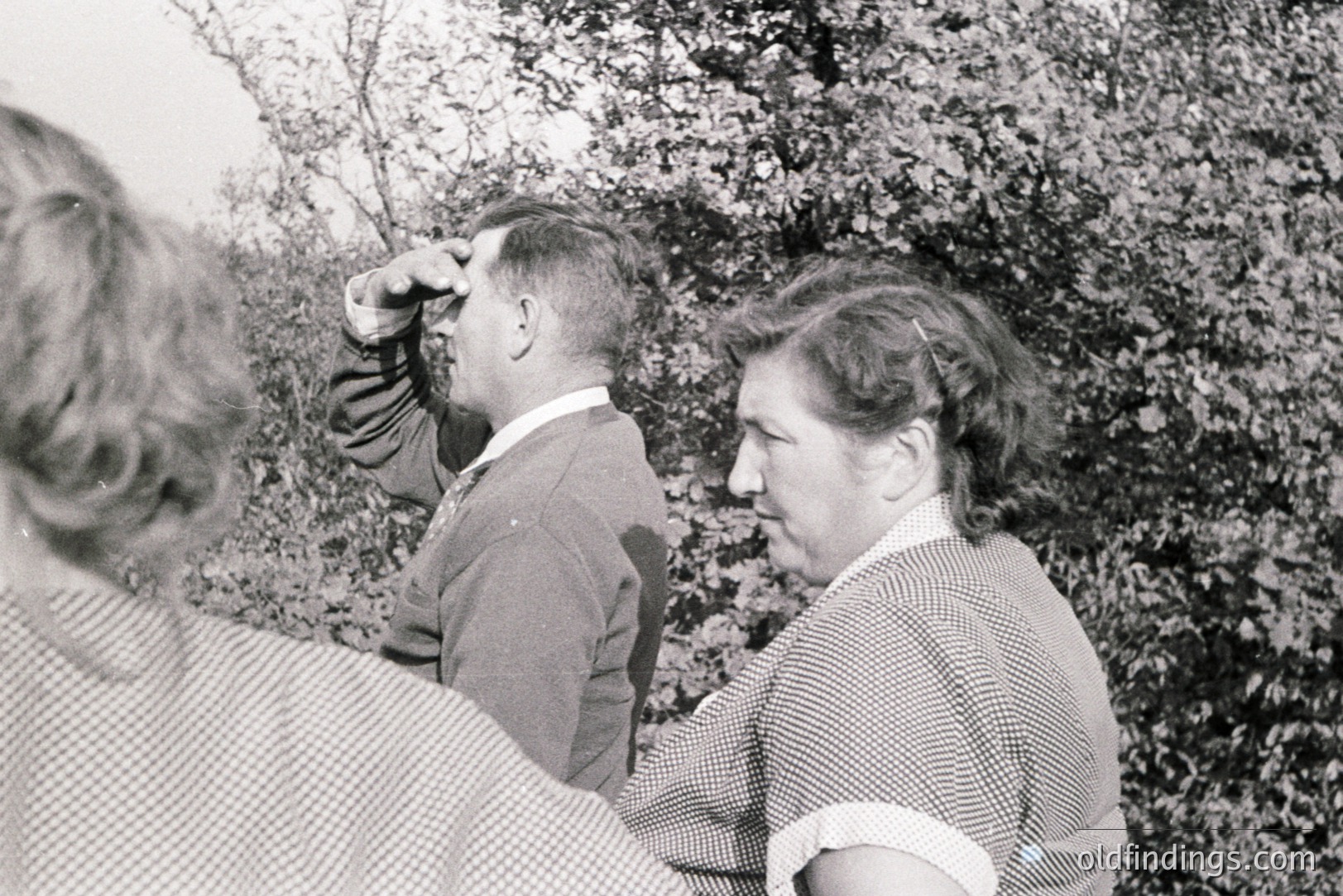 A man in a tweed jacket and collared shirt, peering through what appears to be a telescope, stands next to a woman with a patterned dress and updo. Set against a backdrop of foliage, suggesting a rural or garden setting. Appears to be a candid moment, likely 1950s-1970s.