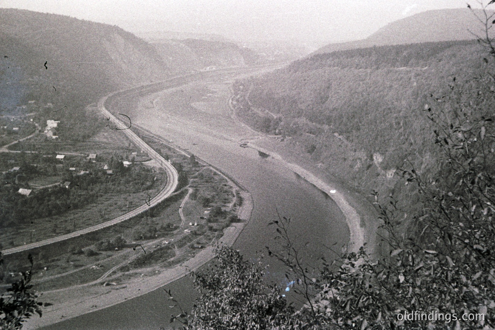Monochrome aerial view depicts a winding river valley, likely a substantial river with barges visible. A curving roadway parallels the river, nestled amongst steep, forested hillsides. Sparse settlement is apparent along the riverbank. Likely post-war era, potentially 1950s or 60s.