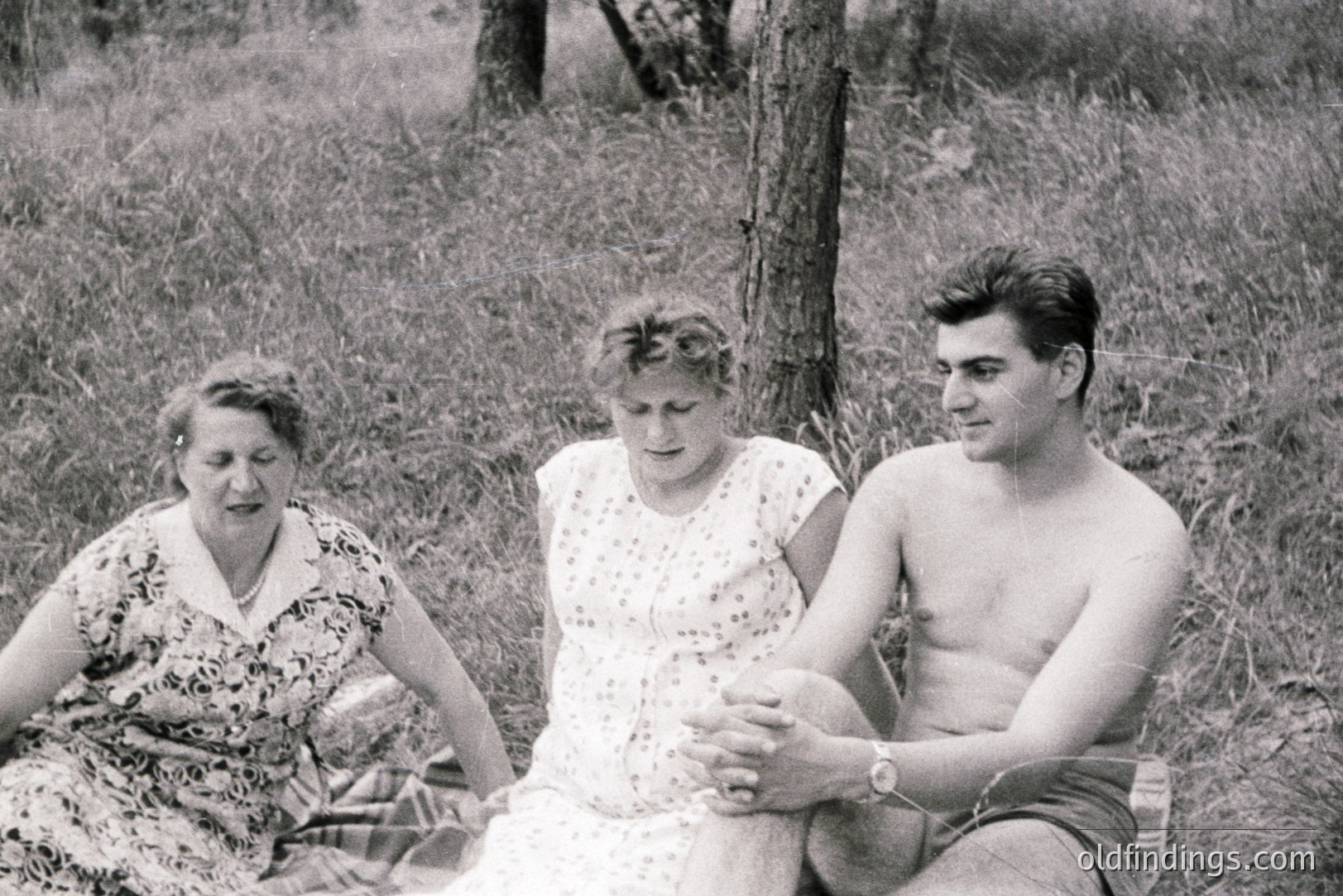 A candid snapshot captures three individuals seated on a blanket amidst tall grass and a blurred treeline. The group appears relaxed; a young man, shirtless and wearing dark shorts, sits cross-legged. Two women, one in a patterned dress and the other in a floral print dress, are beside him. Likely 1960s or 70s, suggesting a casual, outdoor family moment.