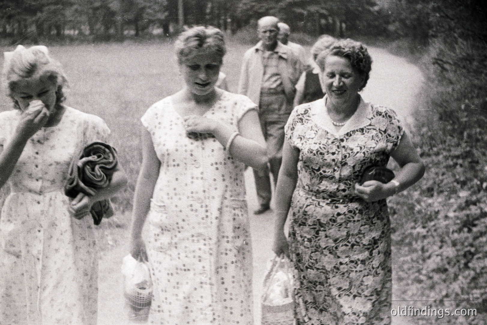 A black and white photo captures a group of people strolling along a path, seemingly in a park setting. Three women in summery dresses lead the way, one dabbing her eyes with a handkerchief. A man and additional figures follow behind. Likely a family outing or social gathering.