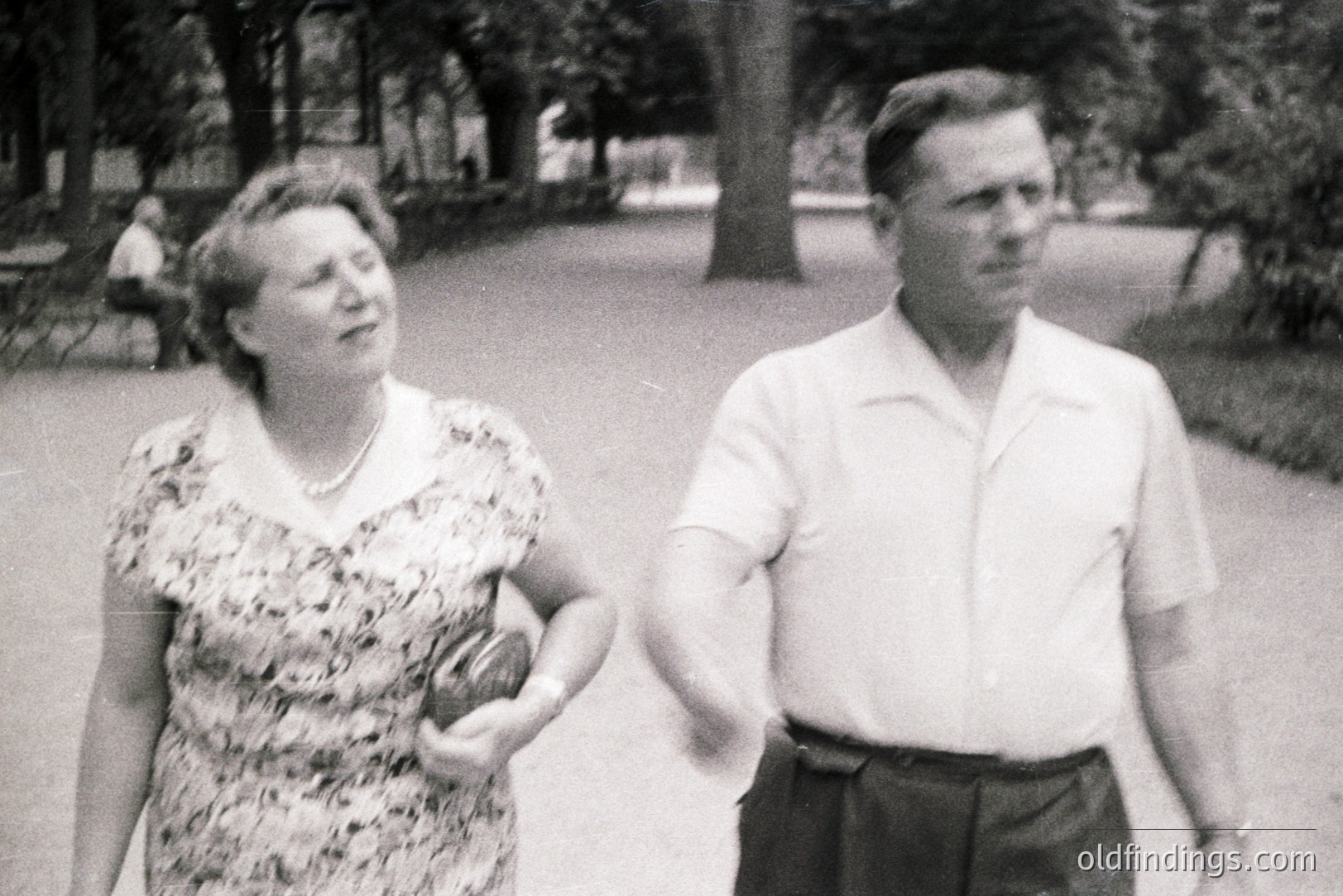 A couple walks along a tree-lined path in a park. The woman wears a patterned dress & pearl necklace; the man, a short-sleeved shirt & pleated shorts. Likely 1960s, possibly European setting. A blurred figure sits on a bench in the background. Image shows vintage aesthetic.
