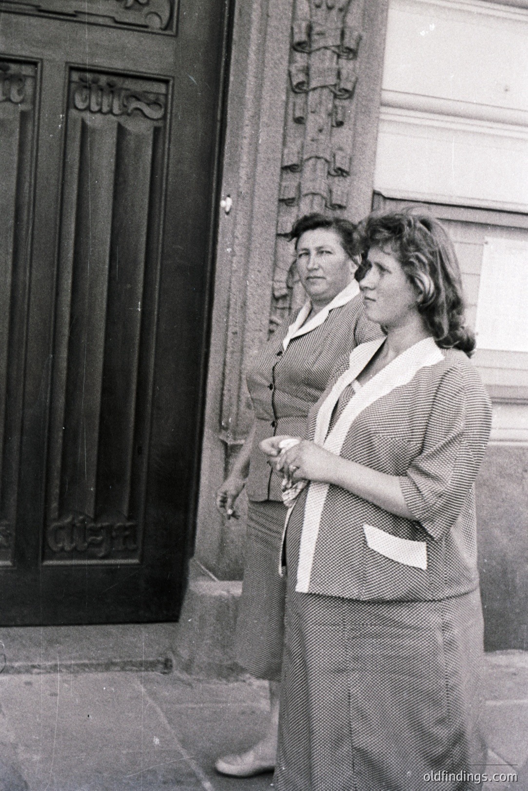 Two women stand outside a building with an ornate door. Both are dressed in matching striped suits with wide collars. Appears to be a candid street portrait, likely 1960s-1970s. The architectural details suggest a European location. A potential stock image for vintage fashion.