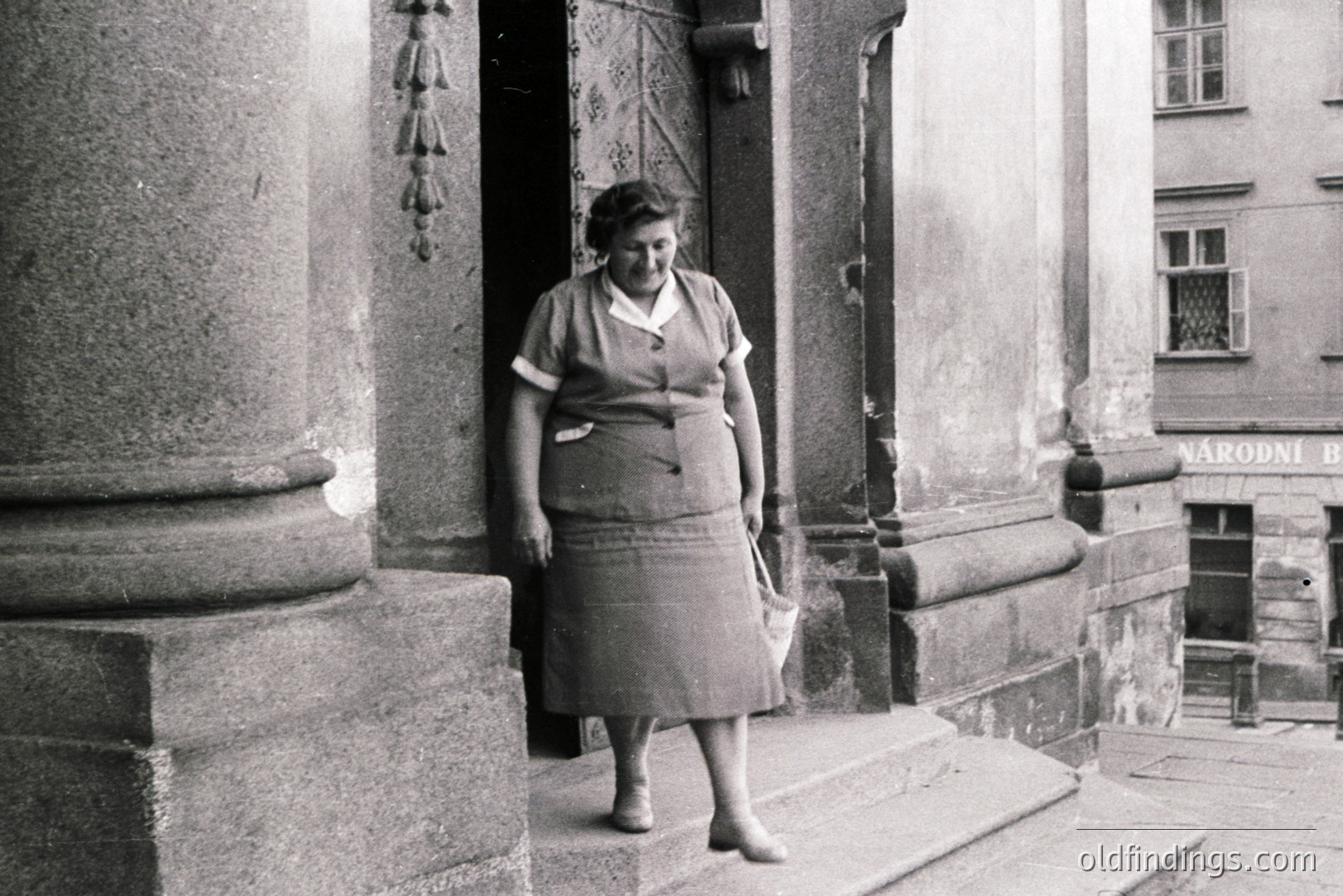 Woman in a tailored suit and skirt descends stone steps, emerging from a building entrance. The architectural details suggest a central European location, likely mid-20th century. Signage in Czech indicates possible Prague. Potential stock photo value for fashion/period research.