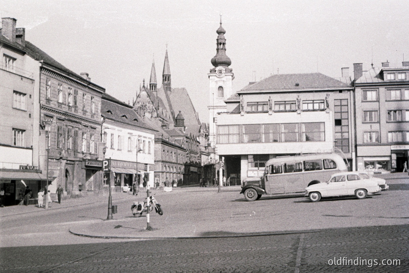 B&W photo depicts a European town square, showcasing a mix of architectural styles. Visible are ornate facades, a church steeple, and early automobiles. Likely post-war, possibly 1950s-1960s. Captures urban life & design of the mid-20th century.