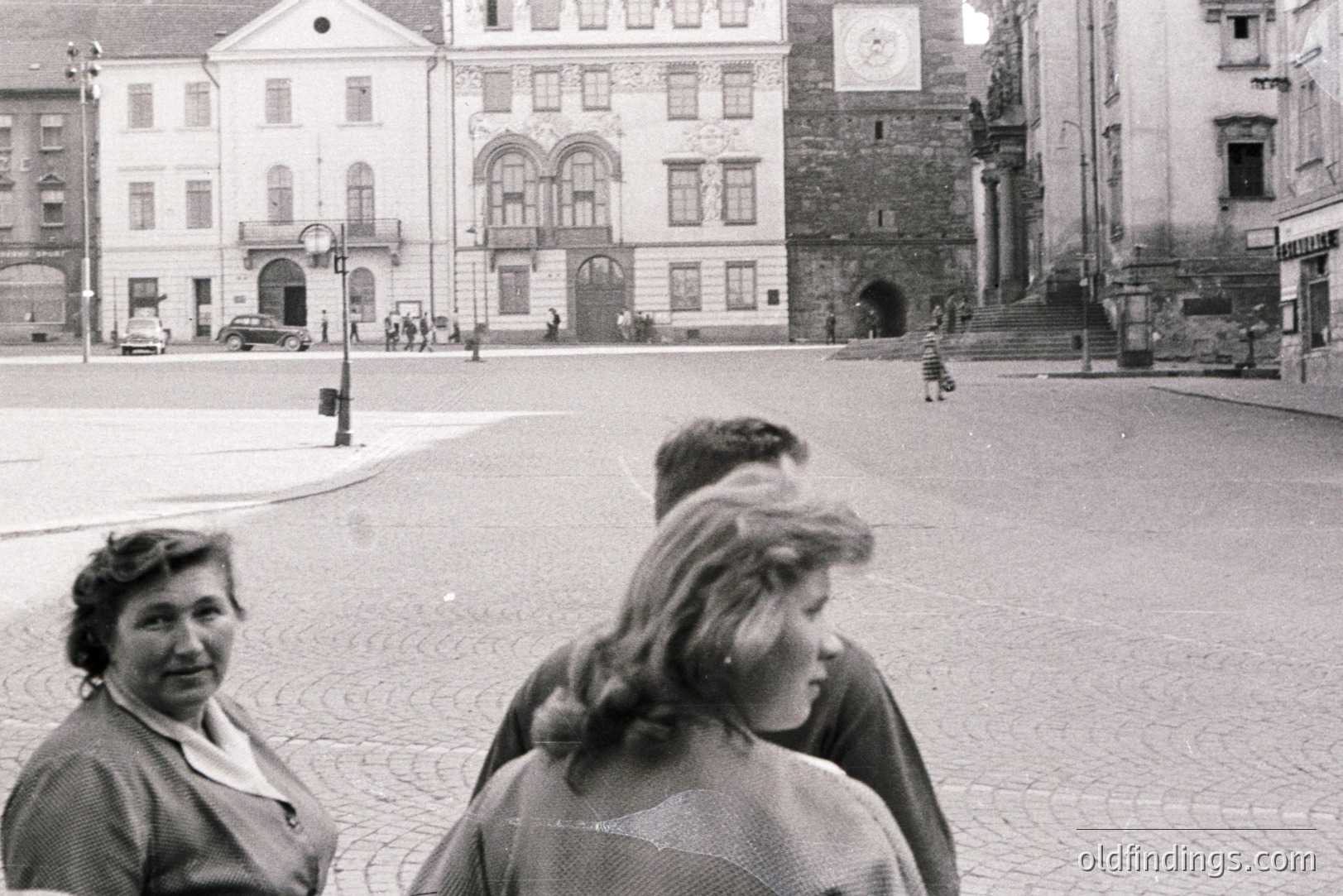 Formal architectural facade dominates a cobblestone square. Three figures are in the foreground, partially obscuring the view. Appears to be a town square with vehicles and pedestrians in the background. Likely a candid street scene, circa 1960s. Useful for historical context or design reference.