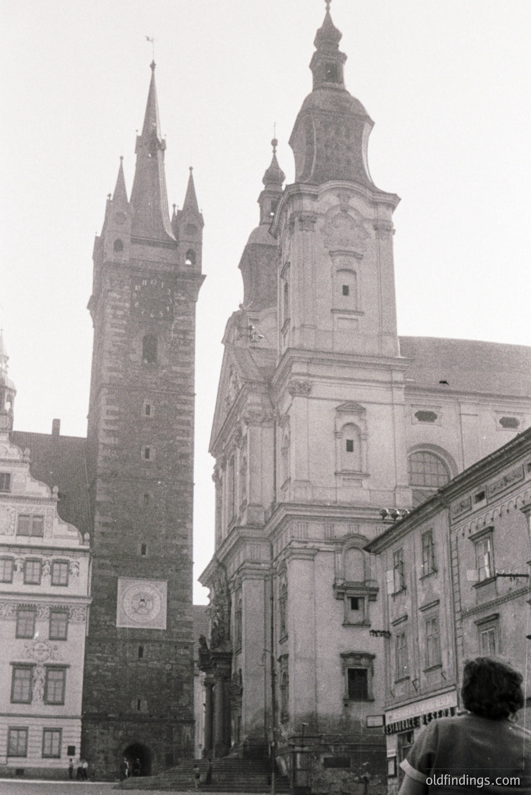 Monumental architecture: A dark stone tower flanks a Baroque church façade with steps leading to the entrance. Note the clock face and intricate detailing. Likely Central Europe, mid-20th century based on photographic style. The partially visible building on the right suggests a town square setting.