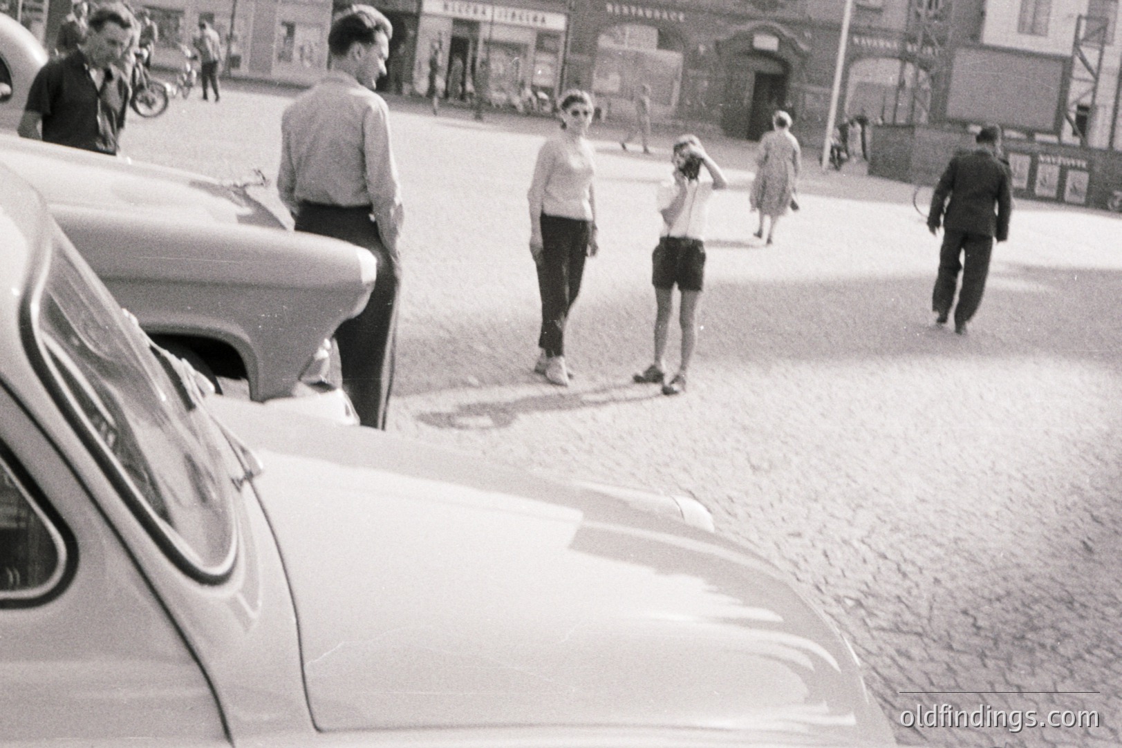 A monochrome street scene captures a group of people in what appears to be a European city. A classic car, possibly a Fiat, dominates the foreground. Three people – two men and a woman – are engaged in conversation. A young girl shielded her eyes. Buildings with signage line the background. Likely 1960s or 1970s.