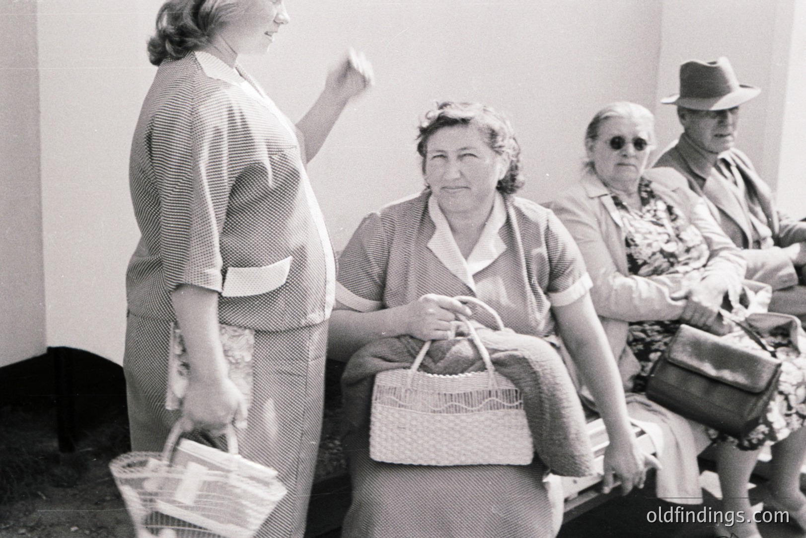 A group gathers, possibly awaiting an event or service. A woman in a striped suit gestures, while others sit with woven bags and hats. Mid-century style clothing & hairstyles suggest the 1950s. Black and white photography captures a candid moment.