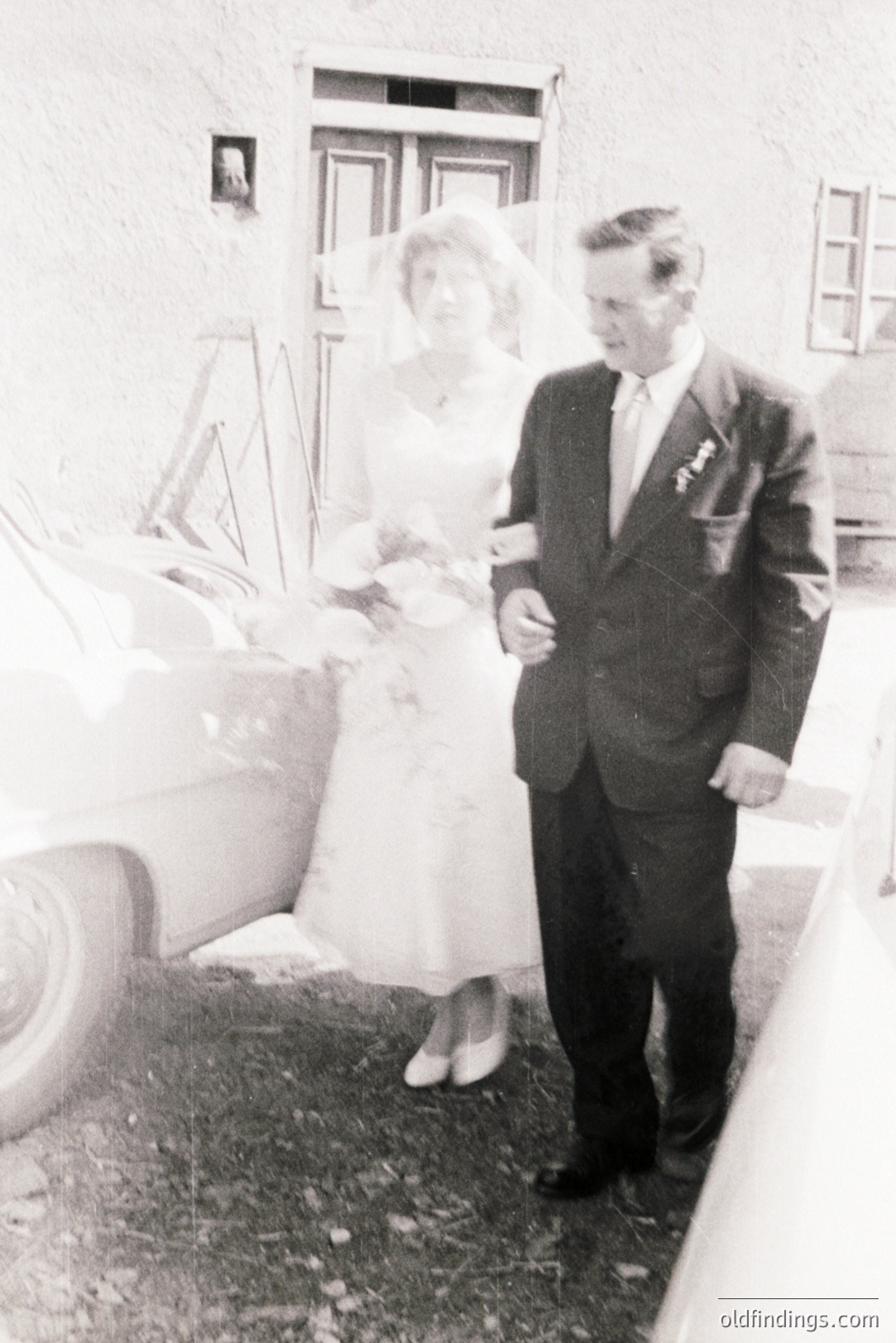 A bride and groom stand beside a classic car, likely post-ceremony. The woman wears a full-skirted wedding dress and veil; the man, a dark suit and tie. Exterior shows a simple building with a ladder & window. Appears to be a candid, informal shot from the 1960s or 70s.
