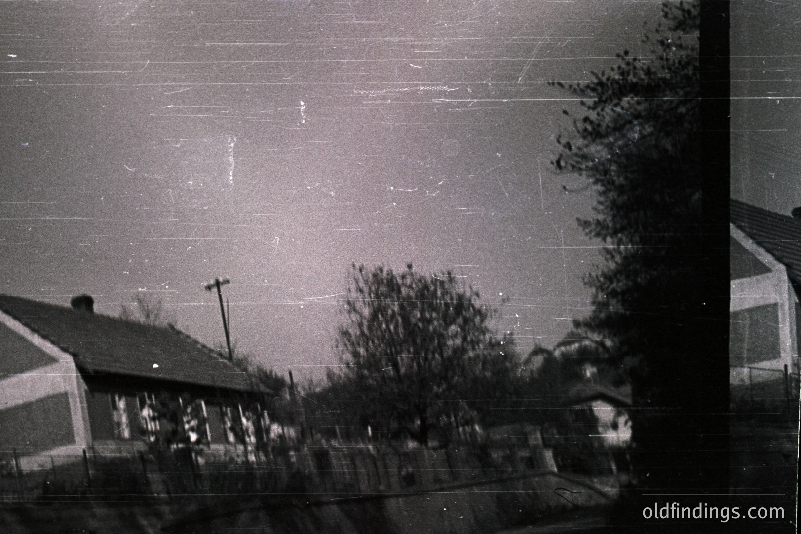 Monochromatic snapshot captures a suburban scene: houses with gable roofs, sparse trees, and a utility pole against a grey sky. Noticeable film grain & scratches suggest age. Likely mid-20th century, possibly 1950s-1970s. Documenting post-war American residential architecture.