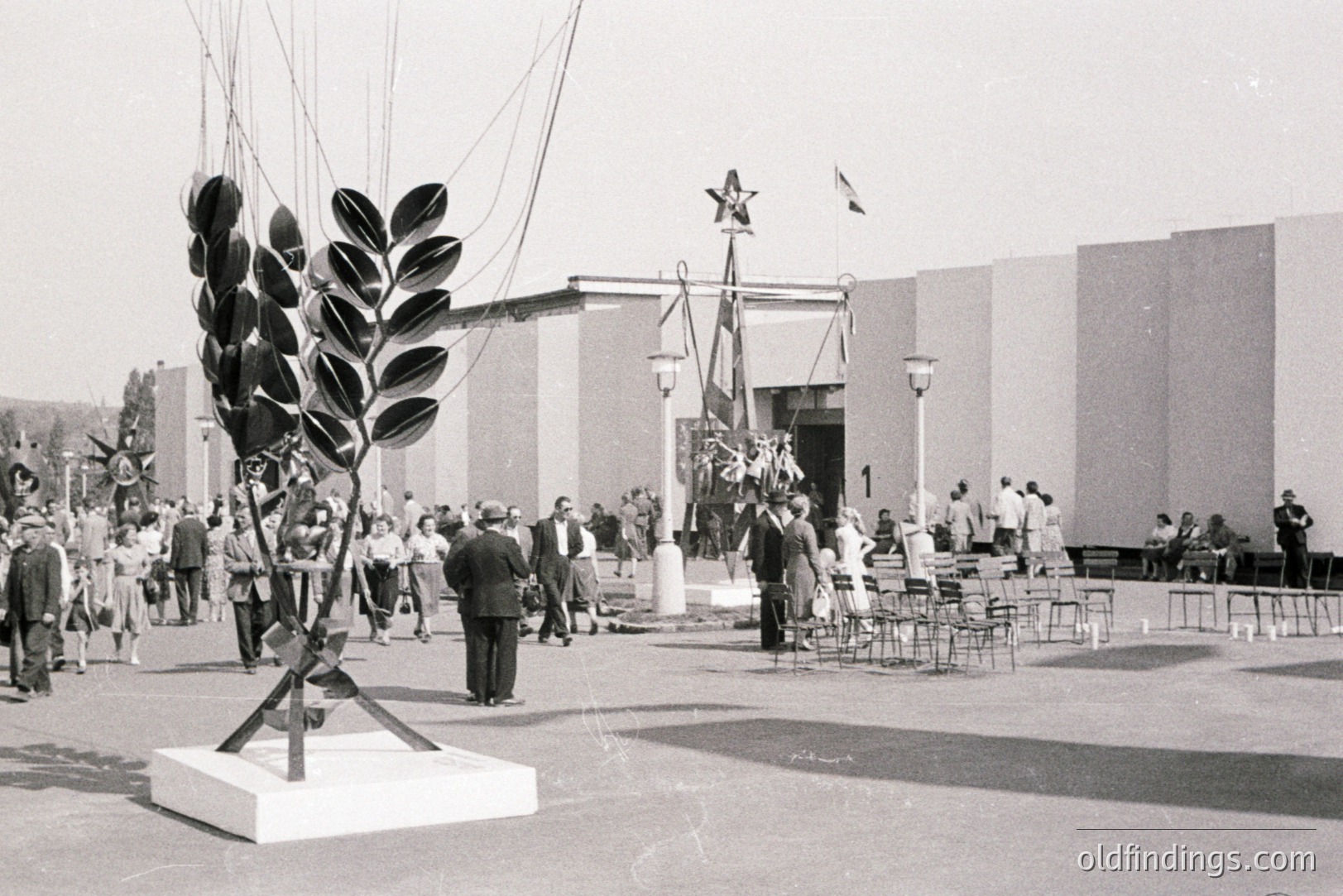 Large-scale, abstract sculpture dominates the foreground of this outdoor scene. A crowd of formally dressed people gathers around flags and a pavilion, suggesting an exposition or national event. Architectural style hints at Soviet influence, circa 1960s. Likely a public display showcasing modern art and national pride.