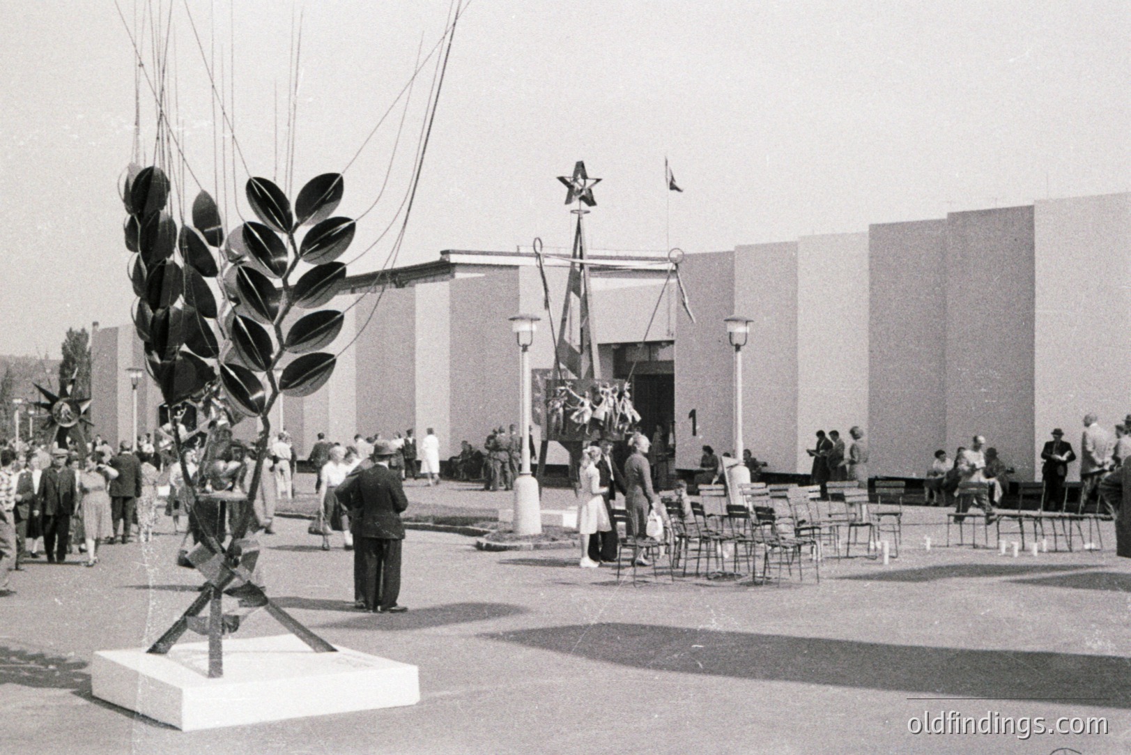 Monumental sculpture of stylized foliage on a plinth dominates this view of a public space, likely an exposition or fair. A stark, modernist building with a central entrance and Soviet-era symbolism (star & flags) is present. Crowds of formally dressed people are visible. Likely 1960s or 70s, Eastern Bloc.