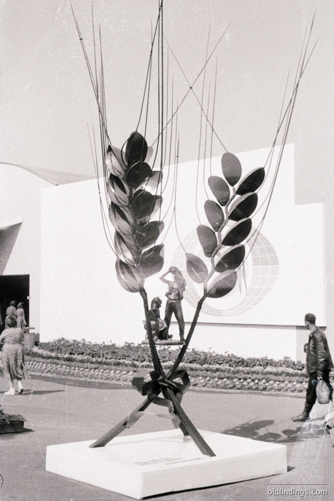 Striking sculpture depicting a wheat stalk with abstract human figures integrated into the design. Likely an exhibition piece from the 1960s or 70s, presented on a white plinth. The artwork references themes of agriculture & humanity. Architectural background suggests a large event or fair.