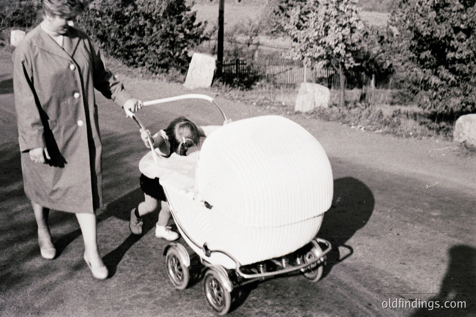 A woman in a long coat pushes a distinctive, rounded baby carriage along a rural road. The carriage features a textured canopy and a close-fitting design, typical of mid-century nursery equipment. A young child is visible inside. Likely a snapshot from the 1950s or 1960s.