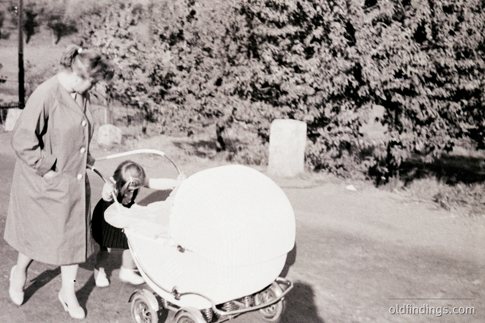 A woman in a knee-length coat pushes a unique, futuristic-looking baby carriage featuring a round basin and chrome accents. The infant is partially obscured within. Appears to be a mid-century, possibly 1950s or 60s, suburban scene. Likely taken in the US or Western Europe. Potential design reference for vintage aesthetics.