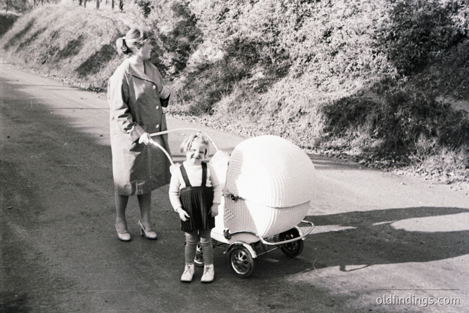 Vintage photograph showing a woman and child in a distinctive wicker baby carriage. The woman wears a long coat & heels; the child, overalls & shoes. Likely 1950s, possibly European setting given carriage style. Carriage's unusual design suggests high-end craftmanship.
