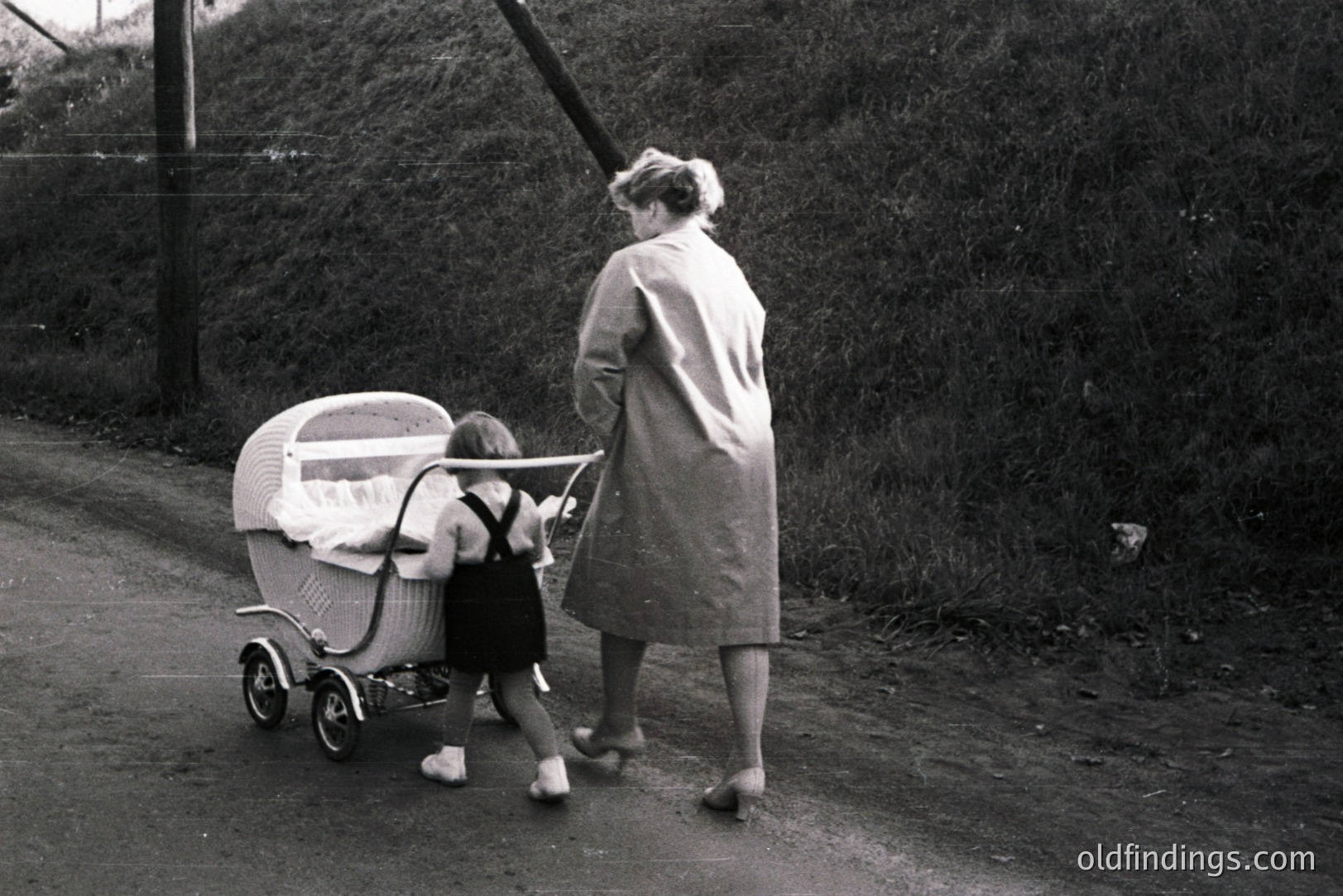 A woman in a trench coat walks along a rural road, pushing an ornate wicker baby carriage. The child, dressed in a dark dress and suspenders, appears to be accompanying her. Likely 1960s, judging by clothing and carriage style. Evokes nostalgia & domestic life. Potential for vintage design reference or historical context use.