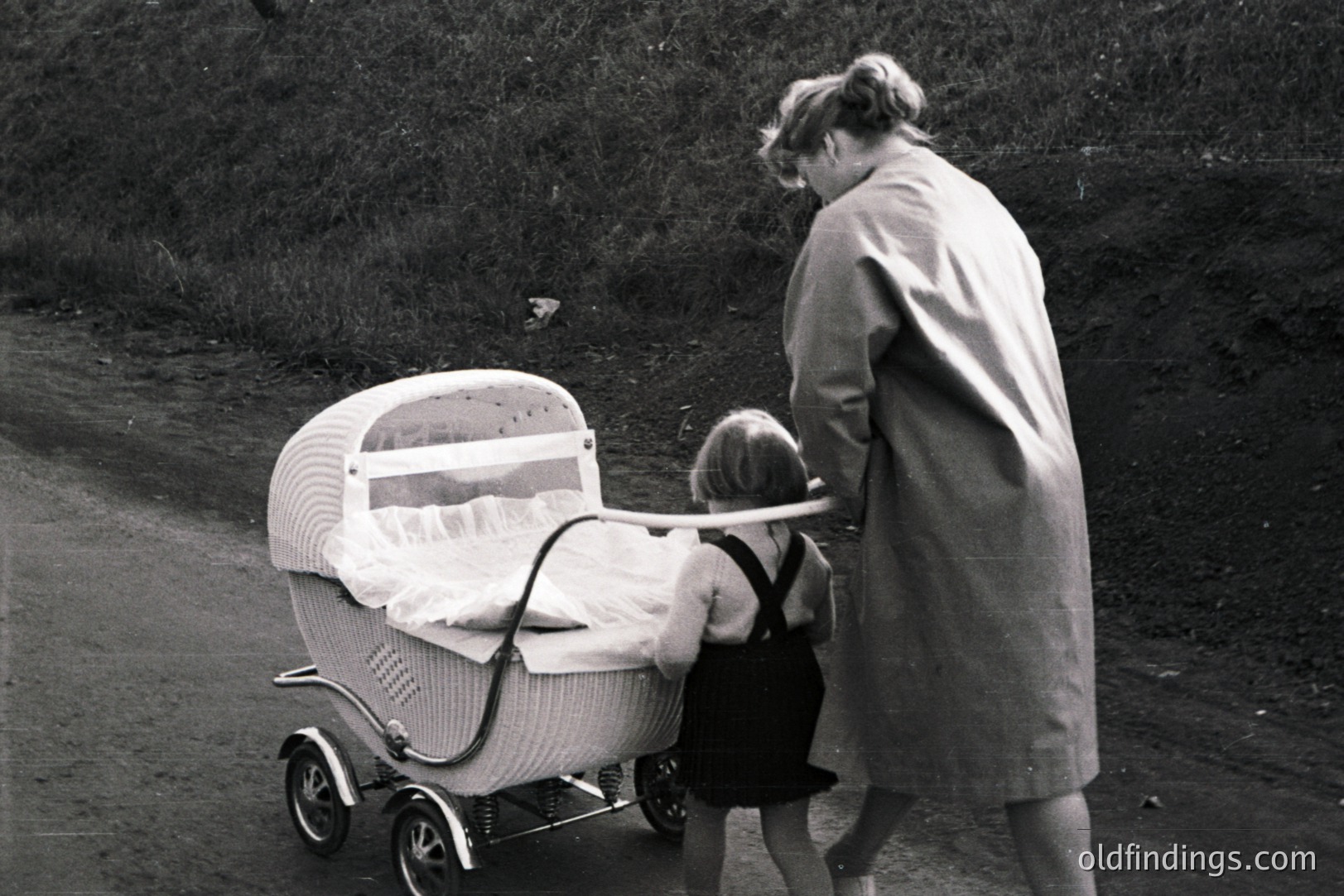 A rear view captures a woman in a long coat pushing a distinctive, woven wicker baby carriage along a gravel road. A child stands nearby, also facing away. The carriage’s design suggests a 1960s or 70s aesthetic. A glimpse of domestic life, likely staged for a catalog or promotional purpose.