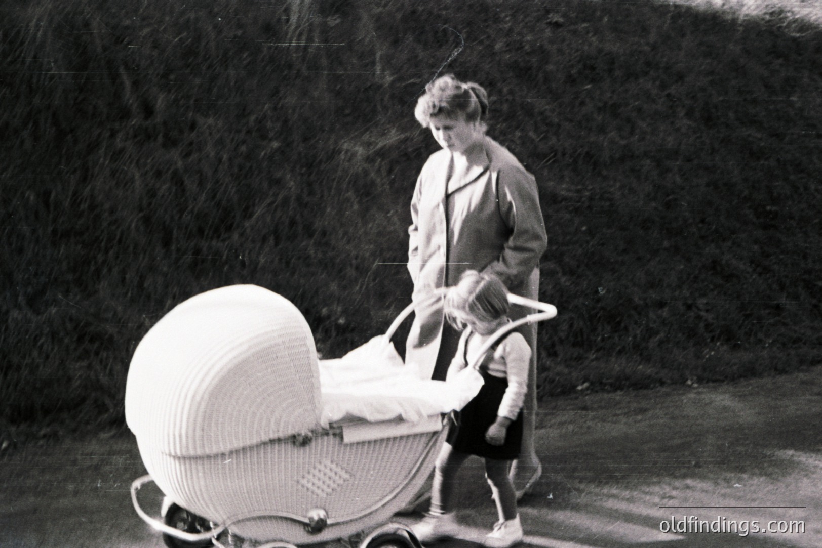 A woman in a long coat pushes a distinctive wicker baby carriage along a paved road. The carriage's curved design and ornate frame suggest a vintage style, possibly 1950s or 60s. The scene has a nostalgic, candid feel. Possibly a stock image for vintage parenting themes.