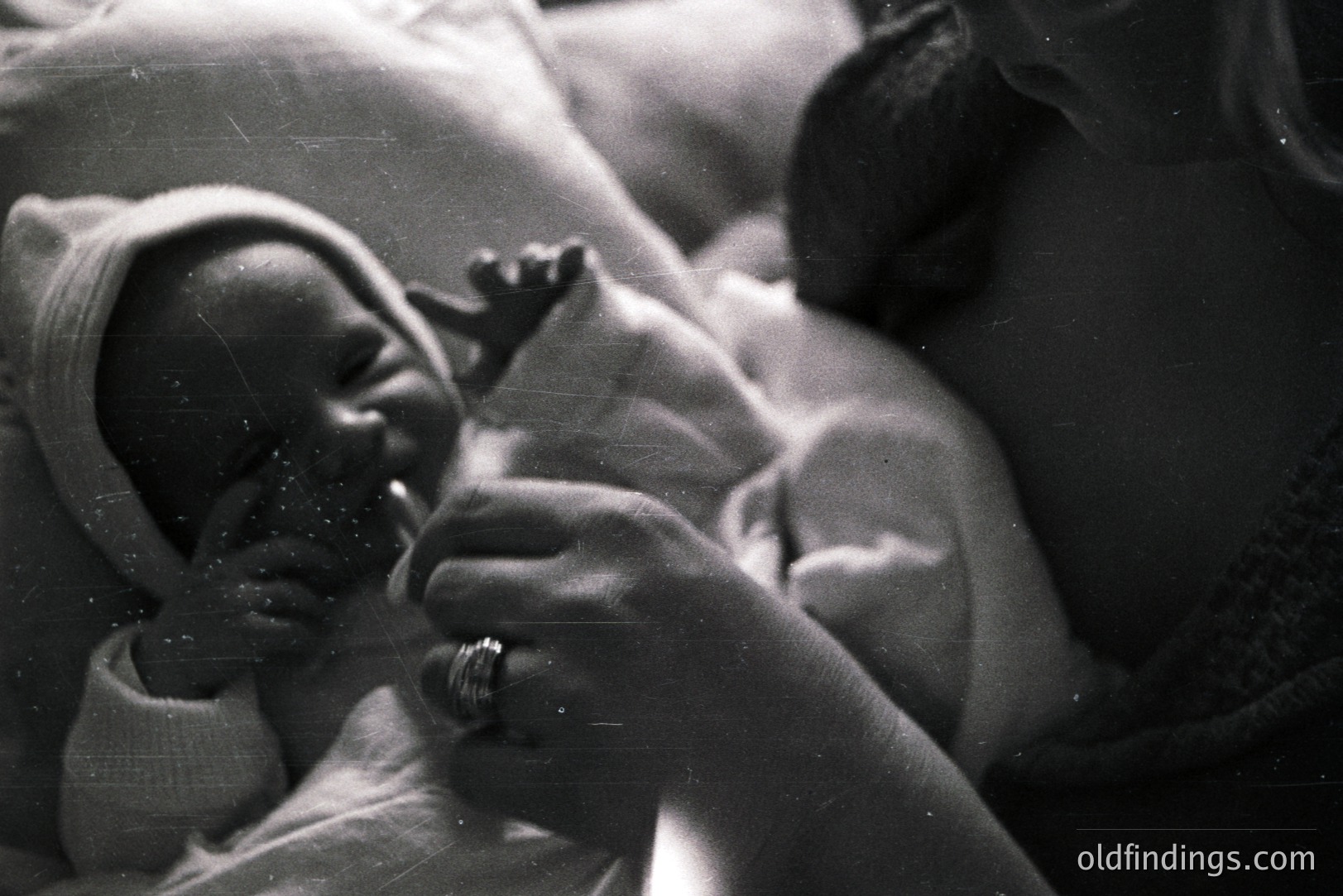 Close-up black and white photograph depicts a newborn infant swaddled in a white blanket, held securely in the arms of an adult. Visible details include the baby's face, a ring on the adult's hand, and textural fabric. Likely a personal or family snapshot from the mid-20th century. Offers potential for design or historical context.