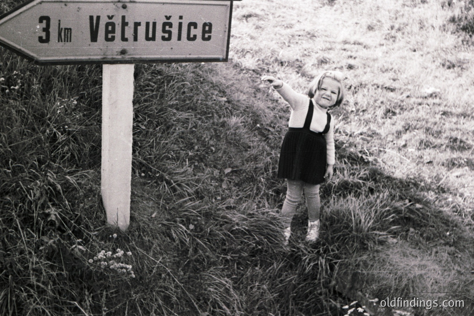 A young girl in a dark dress and bloomers poses next to a weathered signpost indicating "3 km Větrusice." The monochrome image suggests a rural, possibly Eastern European location. The girl's hand gesture and slightly mischievous expression are notable. Likely 1960s-1970s era, documenting childhood and travel.