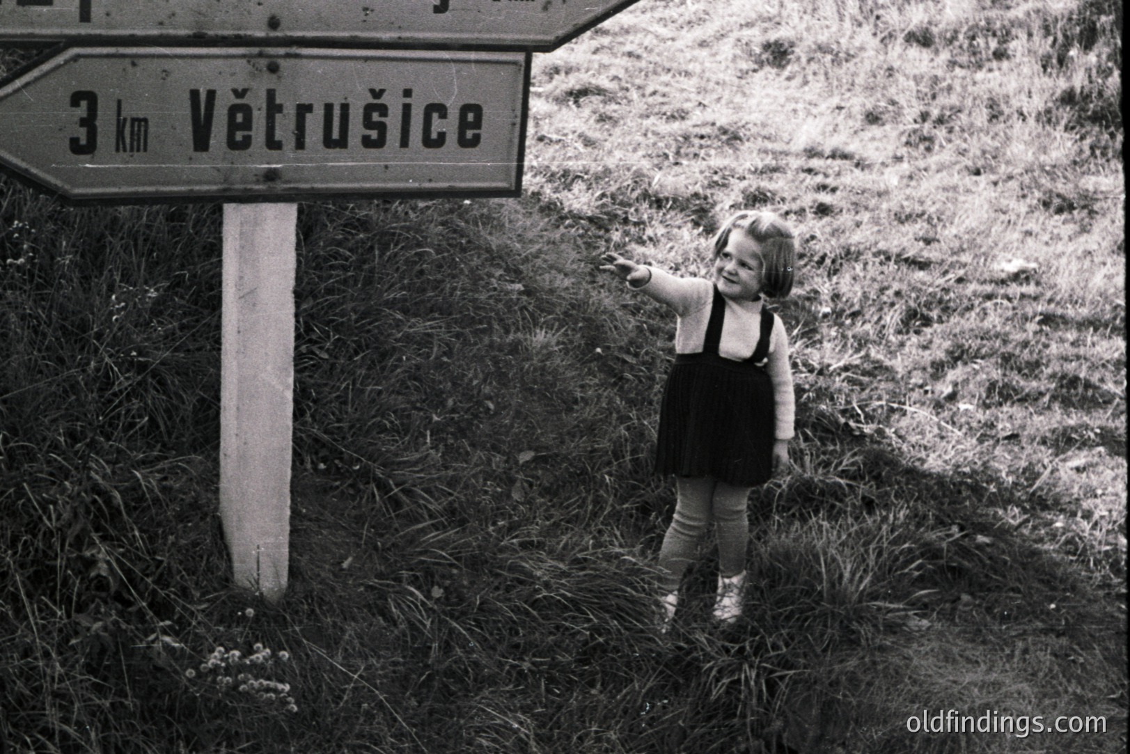 Young girl in a dark dress and tights stands beside a signpost indicating "Větrušice, 3km". The photo, likely from the 1960s or 70s, features a stark contrast in black and white, capturing a candid moment in a rural landscape. A glimpse of a playful childhood.